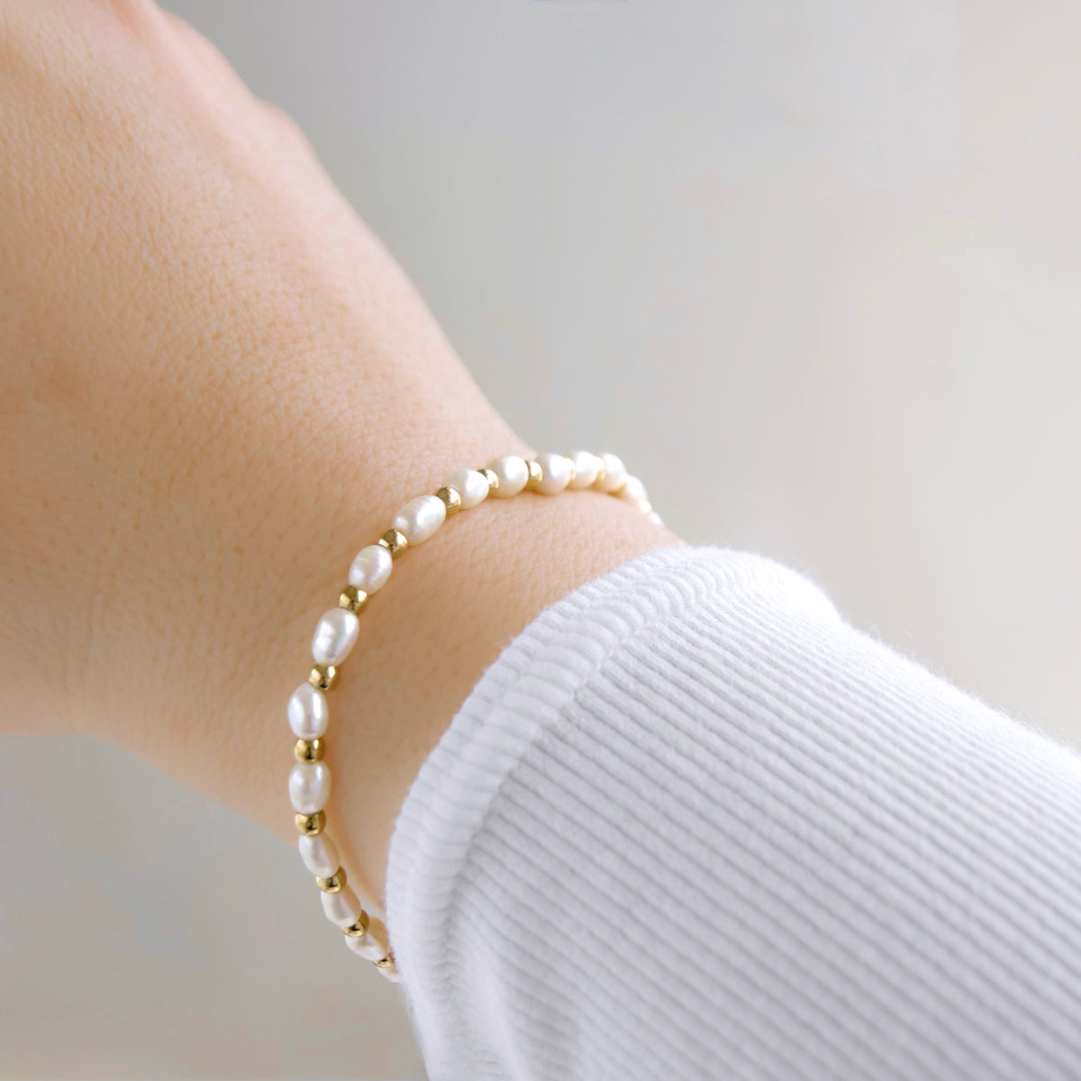 A delicate bracelet with white freshwater pearls and small gold spacer beads, shown worn on a person's wrist over a white ribbed long-sleeve top.
