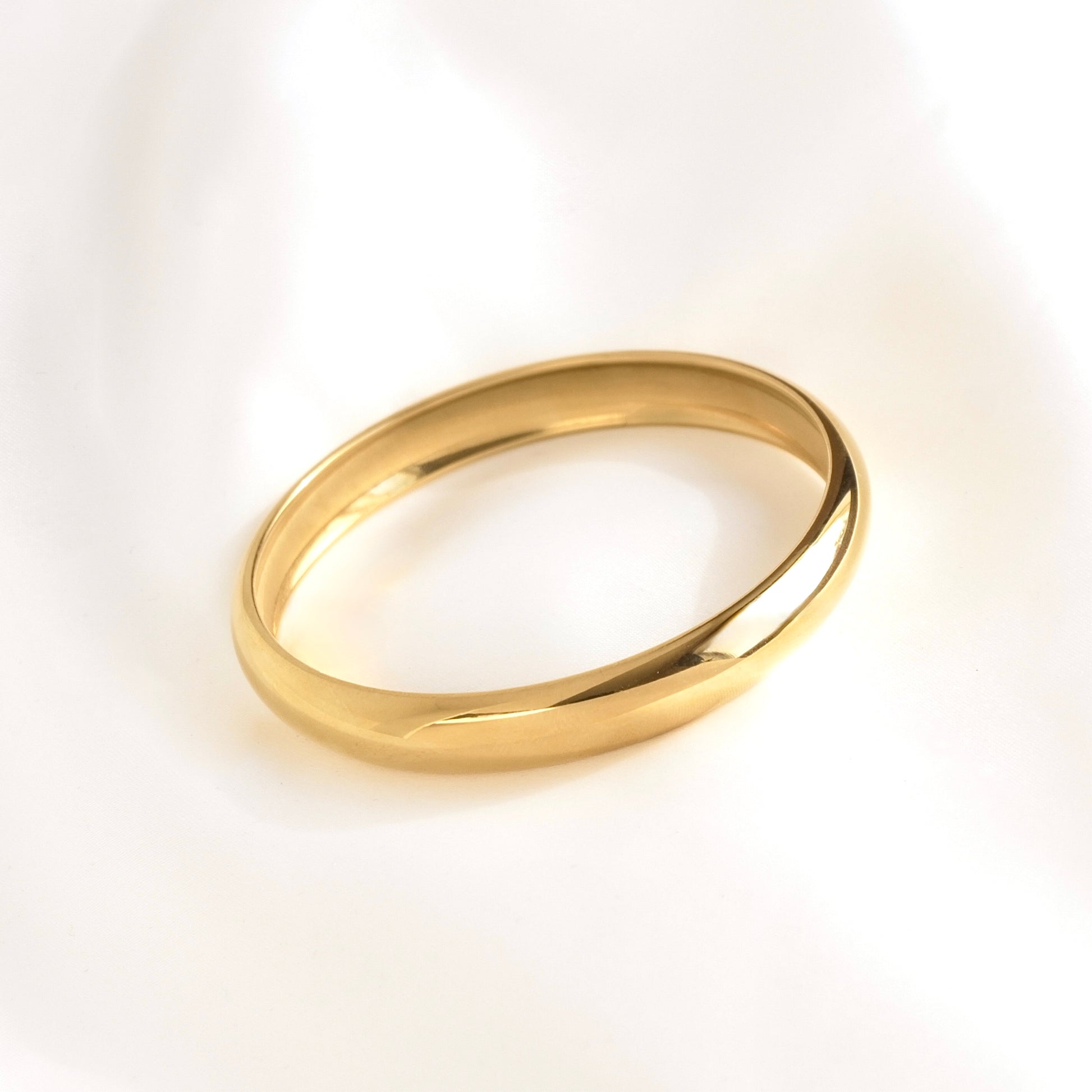 A polished thin gold bangle  with a smooth, rounded profile, resting on a soft white fabric background.