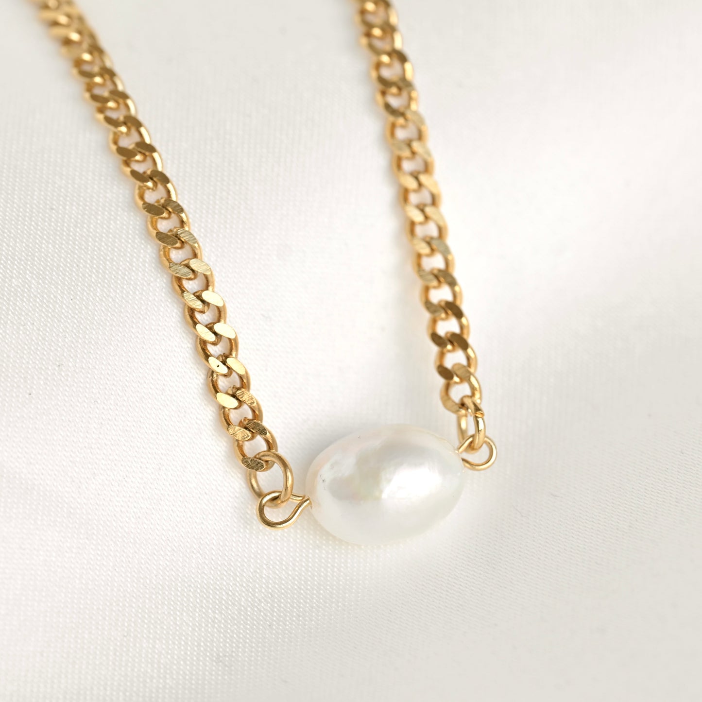 A gold chain necklace featuring a single, irregular white freshwater pearl pendant, displayed on a clean white fabric background.
