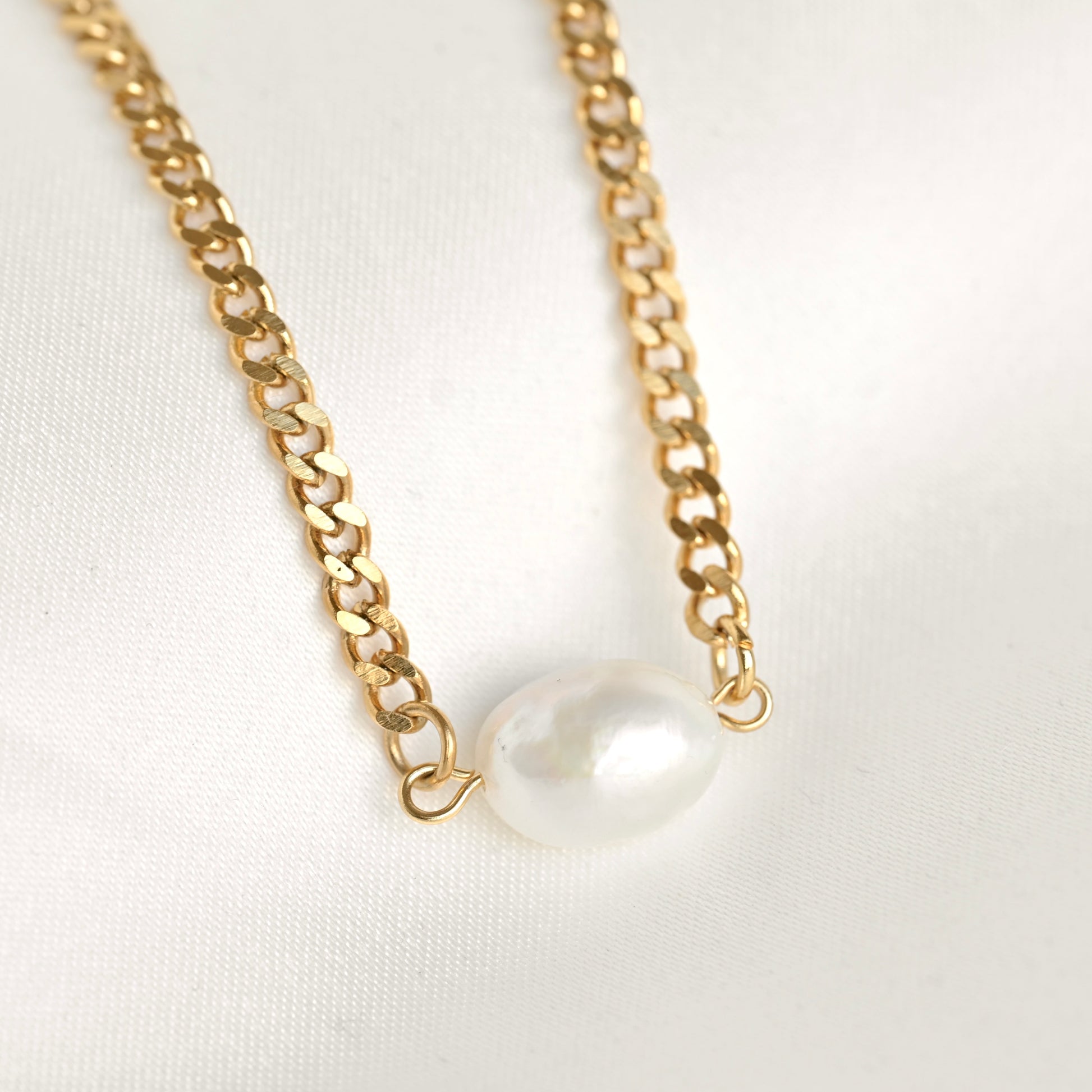 A gold chain necklace featuring a single, irregular white freshwater pearl pendant, displayed on a clean white fabric background.