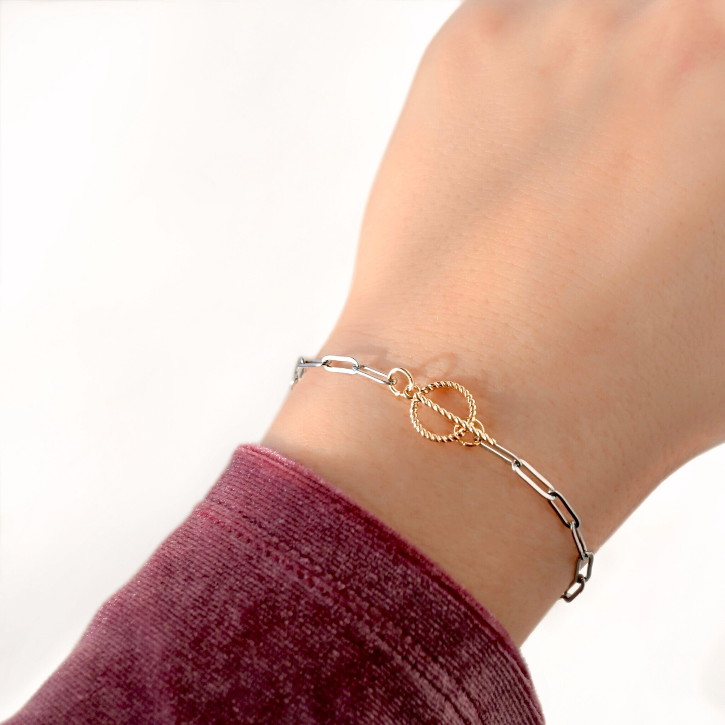 A delicate chain bracelet worn on a wrist, featuring a central gold-toned double-ring motif with one textured circular end and one polished straight end. The bracelet is shown on a person's wrist with a burgundy sleeve visible against a clean white background.