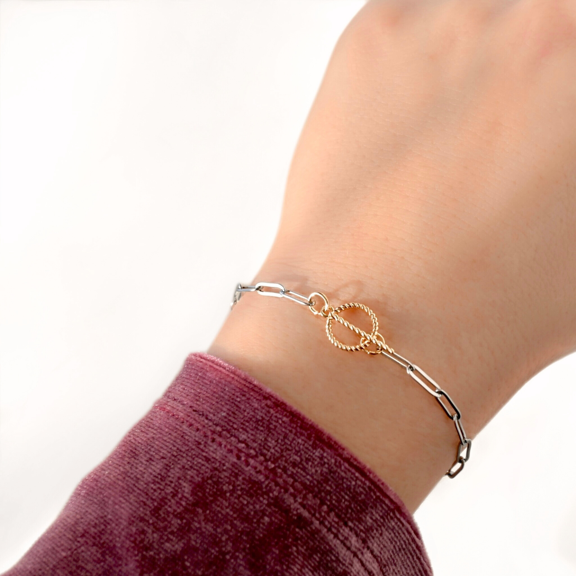 A delicate chain bracelet worn on a wrist, featuring a central gold-toned double-ring motif with one textured circular end and one polished straight end. The bracelet is shown on a person's wrist with a burgundy sleeve visible against a clean white background.