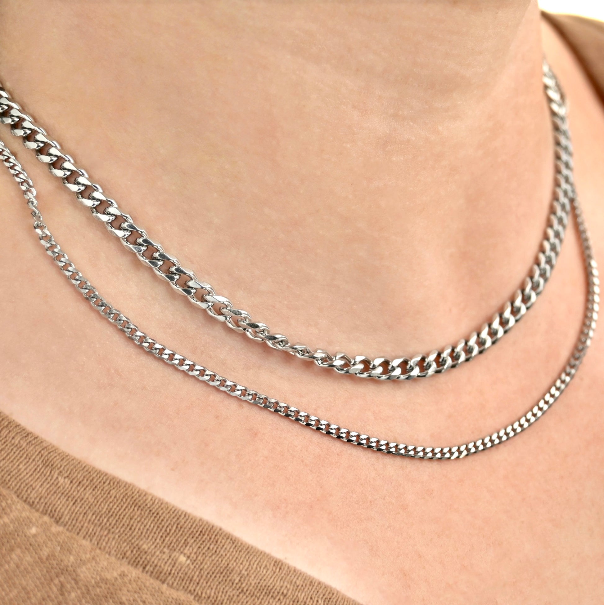 Two silver chain necklaces, one chunky 5mm and one dainty 3mm, on a person's neck against a neutral background