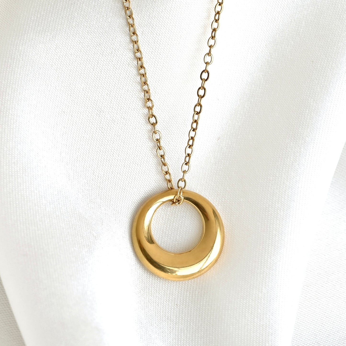 A gold-tone necklace with a smooth, polished tapered circle pendant on a delicate chain, displayed on a white fabric background.