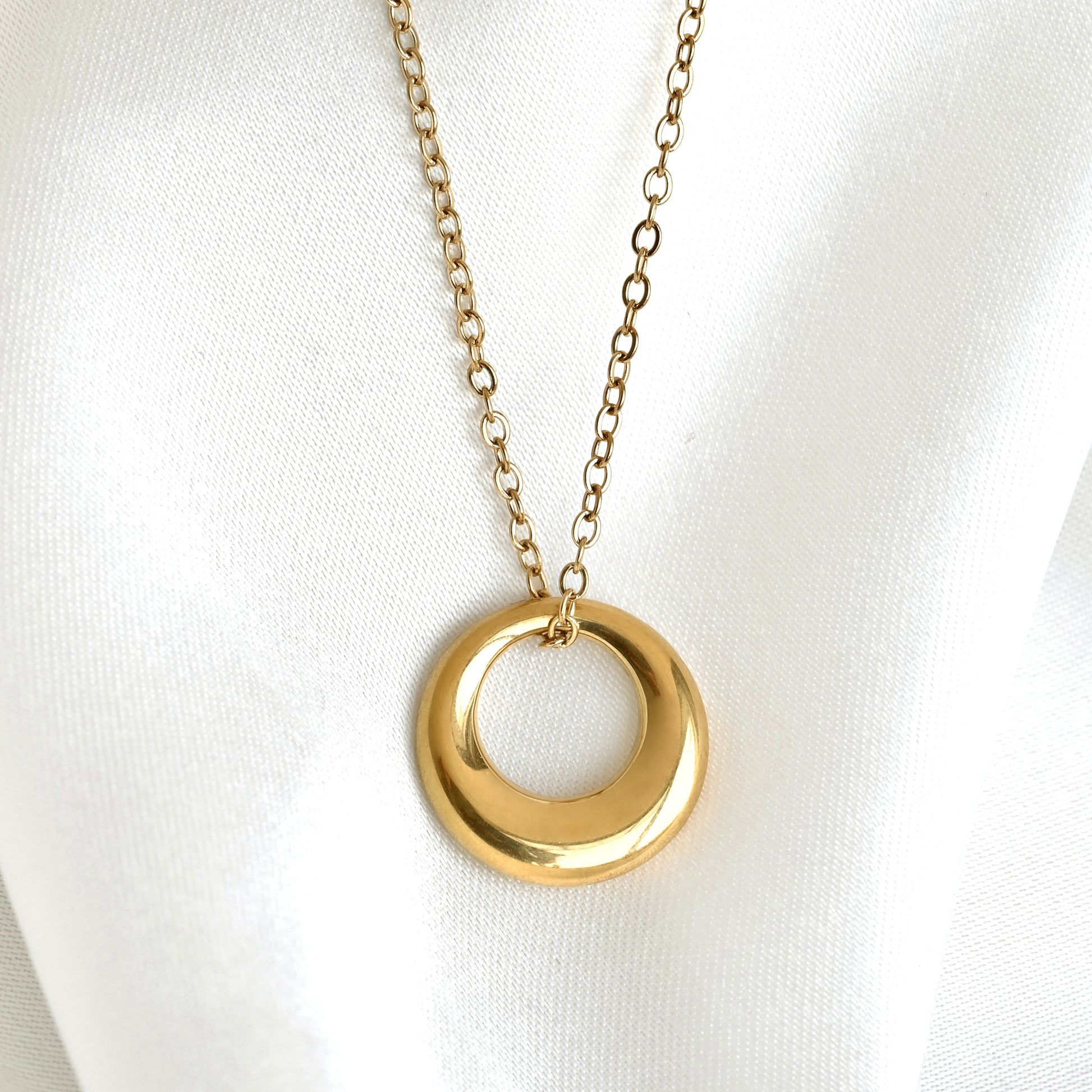 A gold-tone necklace with a smooth, polished tapered circle pendant on a delicate chain, displayed on a white fabric background.