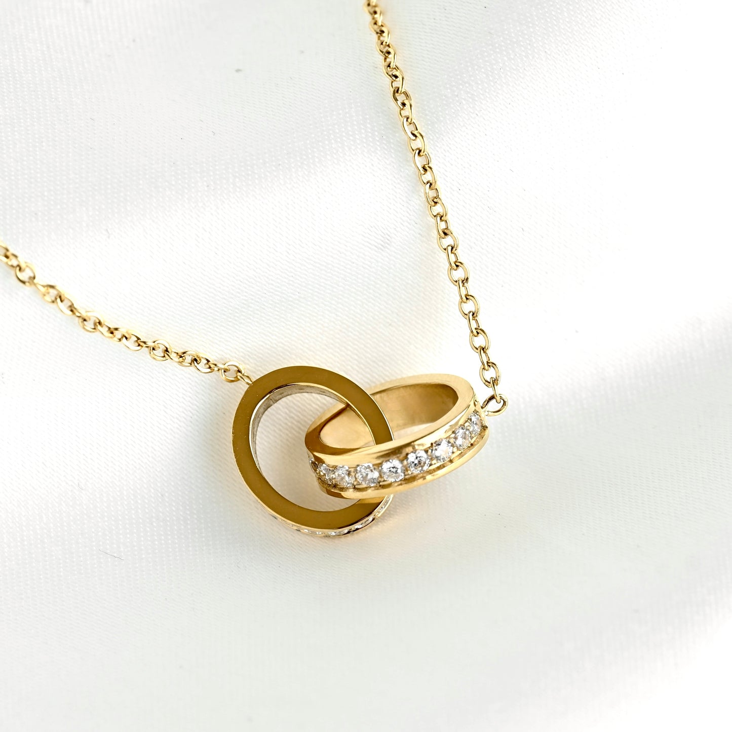 A gold chain necklace with a pendant consisting of two interlocking rings, one plain and one paved with small clear stones, displayed on a white textured fabric background.