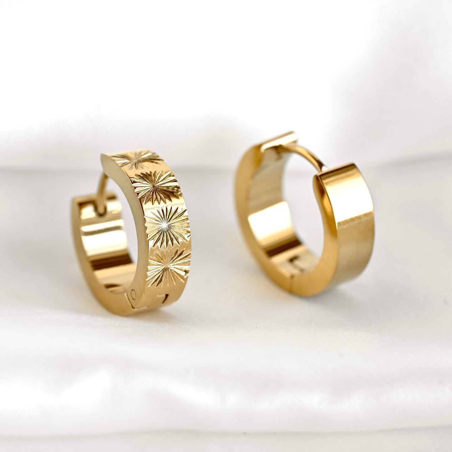 A pair of gold huggie hoop earrings with a sunburst star pattern engraved on the front and high-polish smooth finish on the back, displayed on a soft white fabric background.