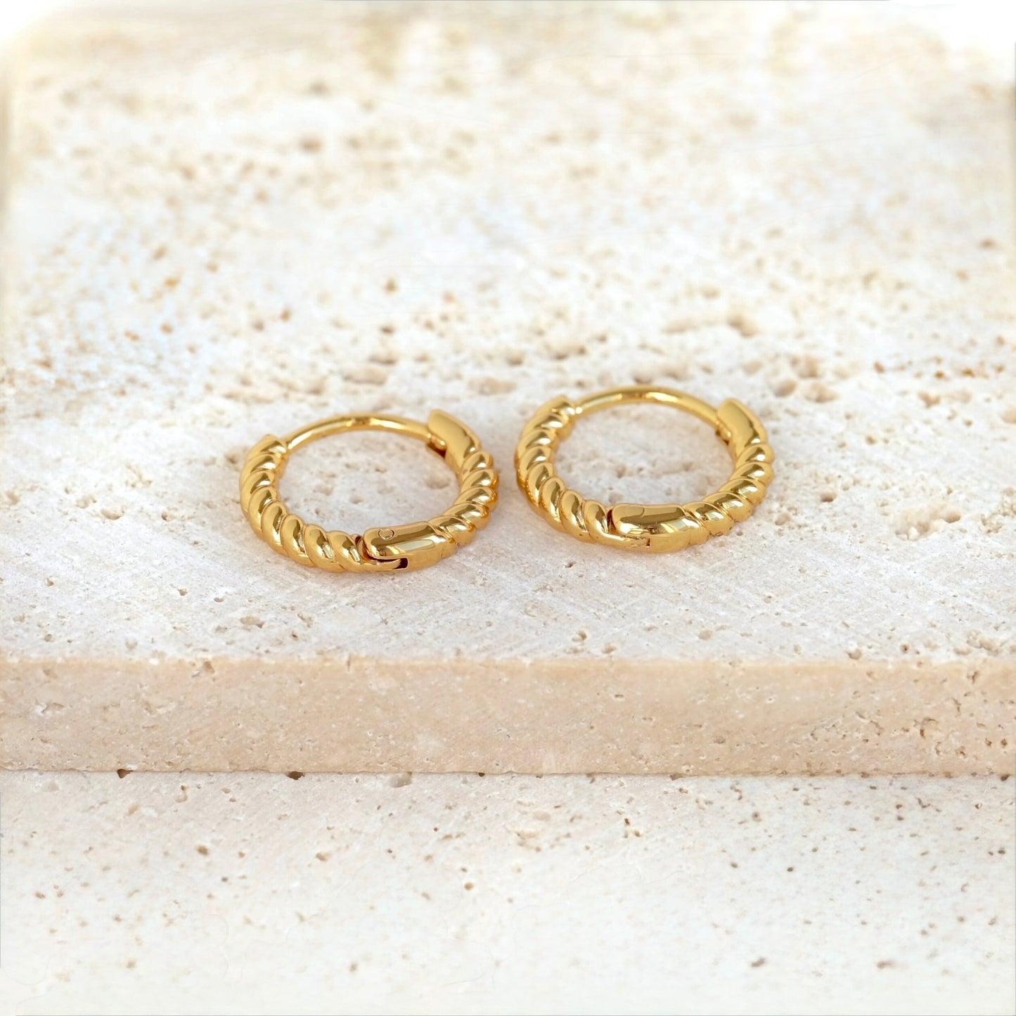 A pair of small gold huggie hoop earrings with a twisted rope texture, resting on a light beige stone slab.
