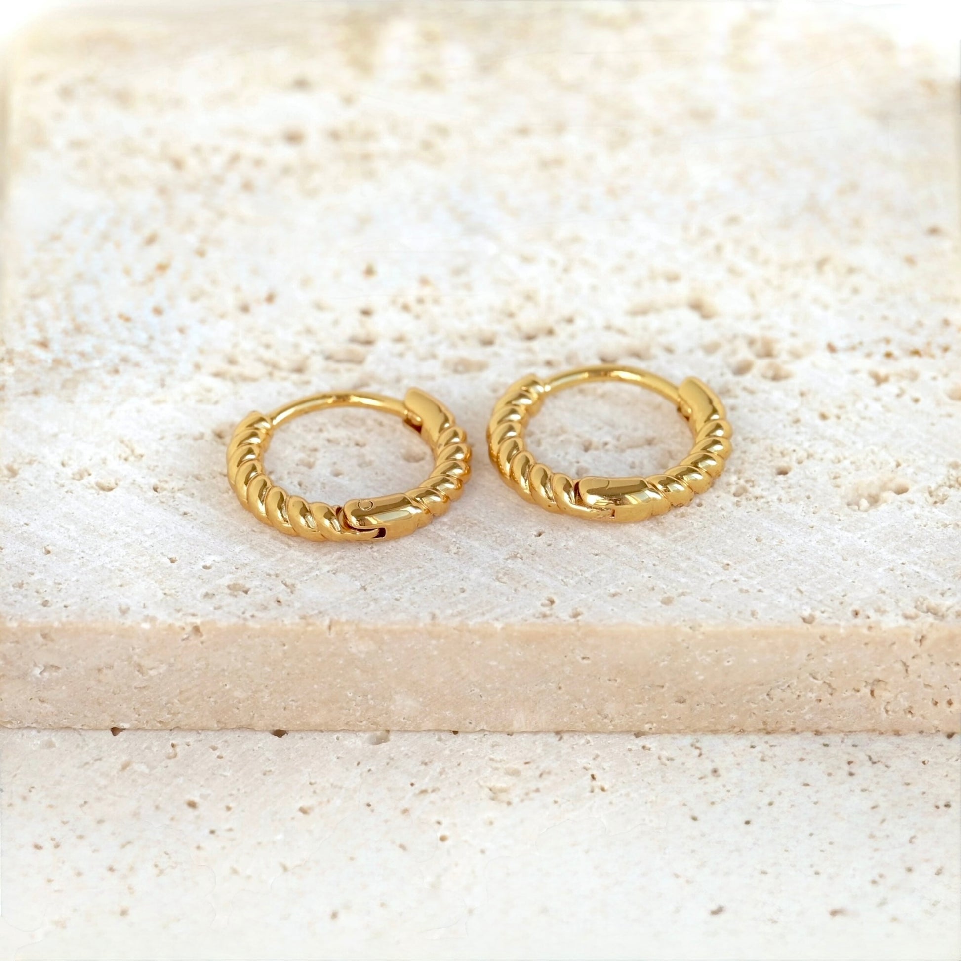 A pair of small gold huggie hoop earrings with a twisted rope texture, resting on a light beige stone slab.