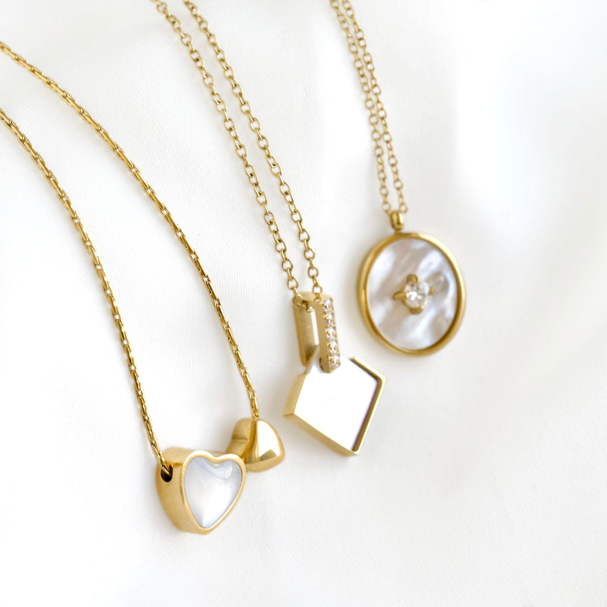 Three gold necklaces with mother-of-pearl pendants arranged on a white fabric background. The necklaces feature heart, square, and oval pendants with a small clear stone in one.