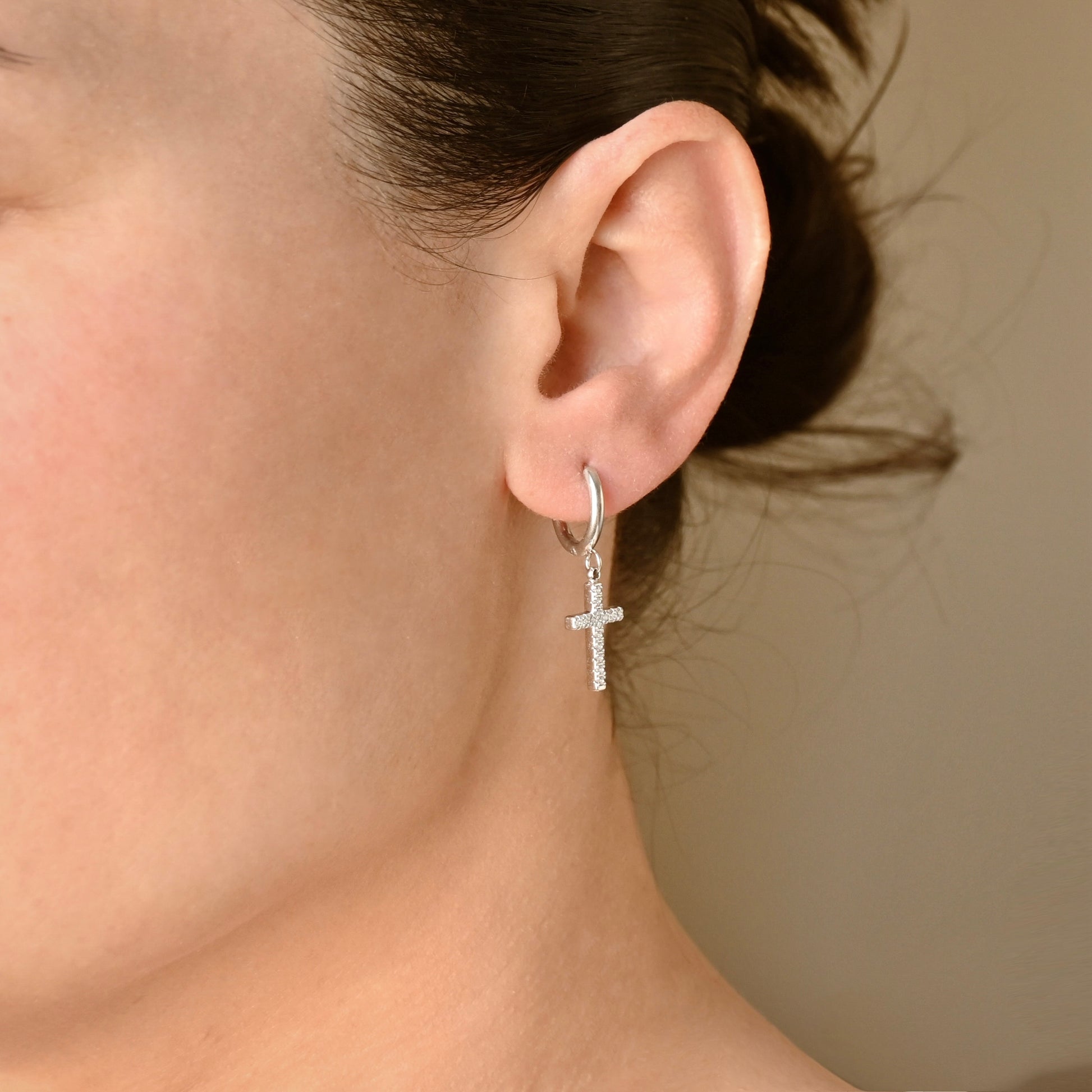 A close-up of a person's ear wearing a small silver hoop earring with a diamond cross pendant, shown against a plain beige background.
