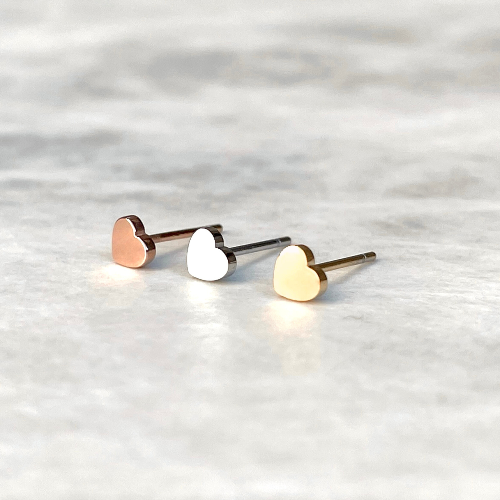 Front view of mini heart-shaped stud earrings in rose gold, silver, and gold on a light gray background