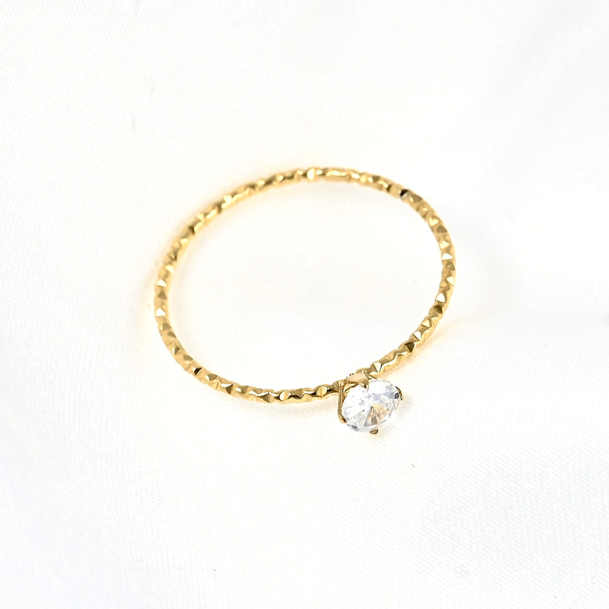 A textured yellow gold ring with a single clear round gemstone, photographed on a white fabric background.