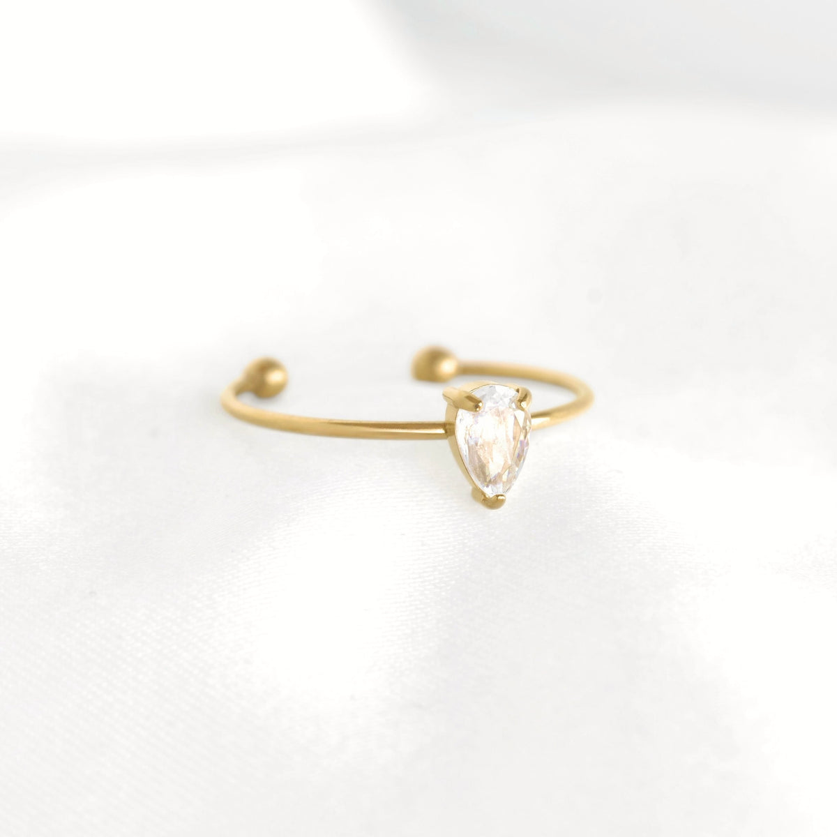 A delicate gold open-ended ring featuring a small pear-cut clear gemstone in a prong setting, resting on a soft white fabric surface.