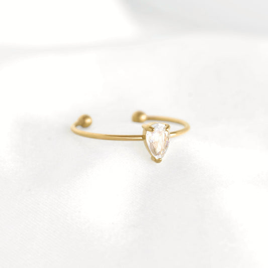 A delicate gold open-ended ring featuring a small pear-cut clear gemstone in a prong setting, resting on a soft white fabric surface.