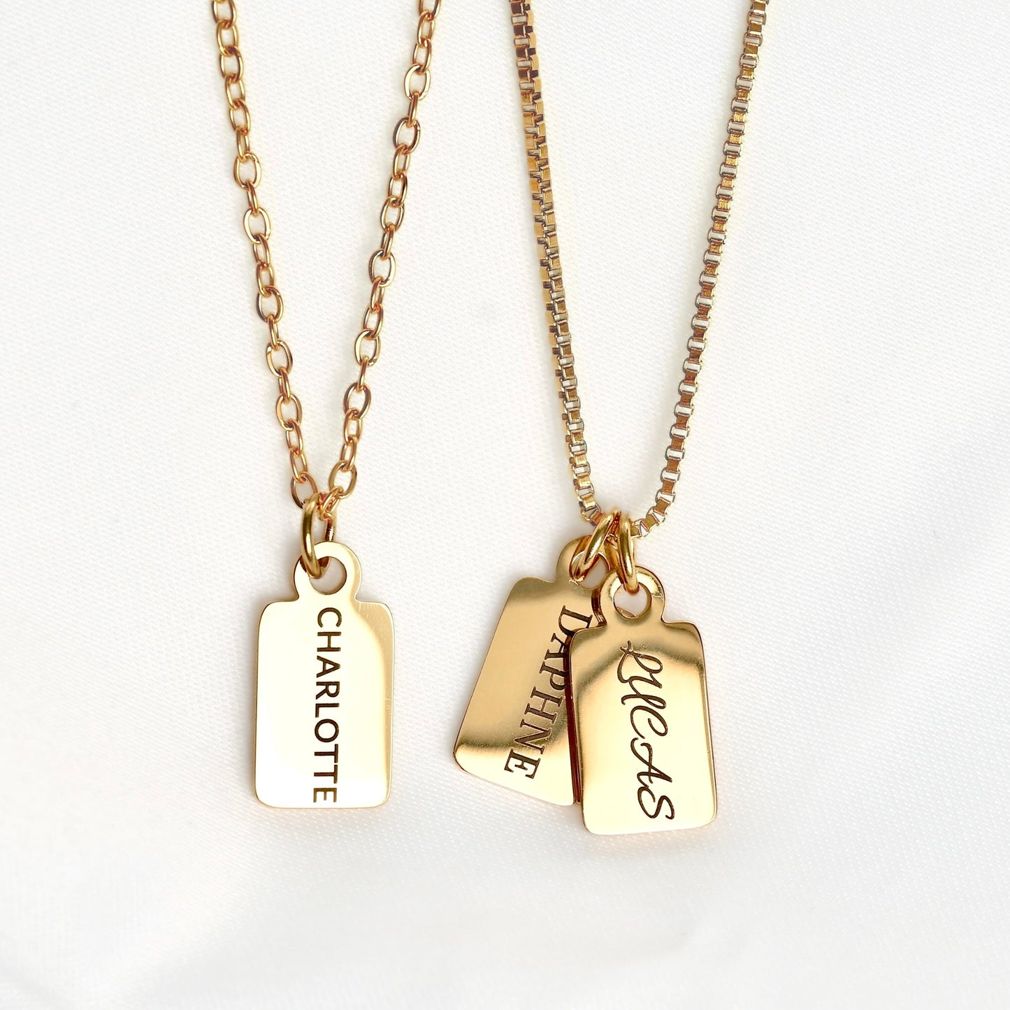 Two gold-tone personalized necklaces with rectangular hanging tags on chains, displayed on a white fabric background. One tag is engraved with the name 'CHARLOTTE' and the other with 'DAPHNE' and 'LUCAS'.