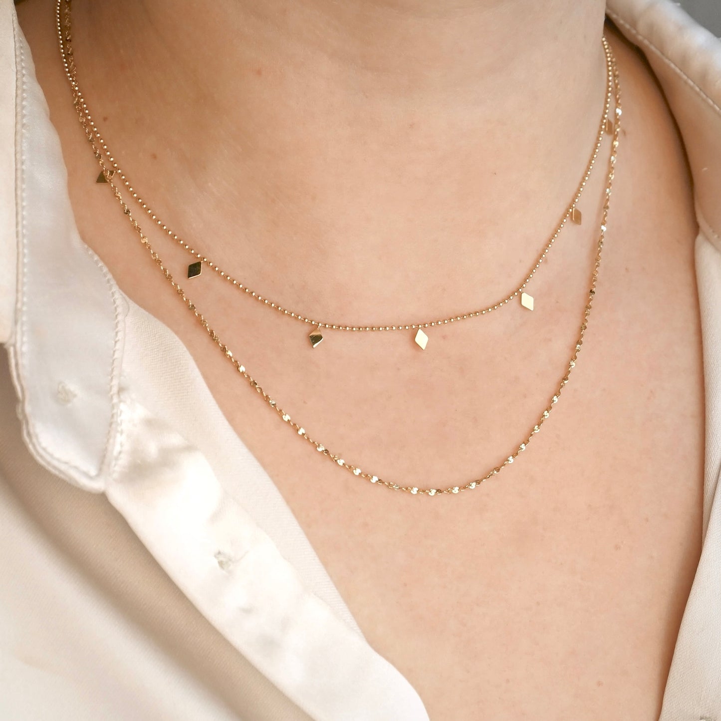 Angled front view of a person wearing a layered gold necklace set consisting of a long twisted sparkle chain and a shorter ball chain with rhombus shaped beads stationed along.