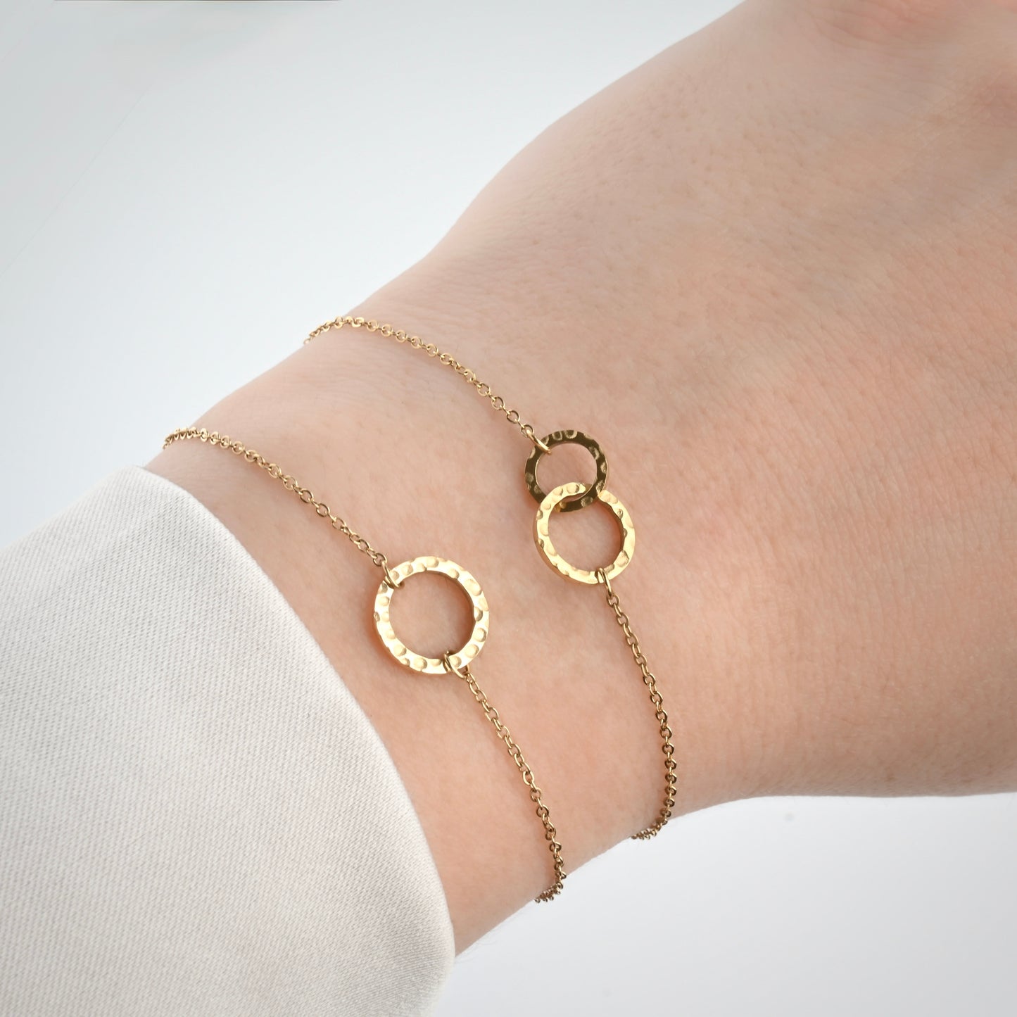 Two gold chain bracelets with hammered circle links on a wrist, one with a single medium circle, one with two different sized interlocking circle charms.