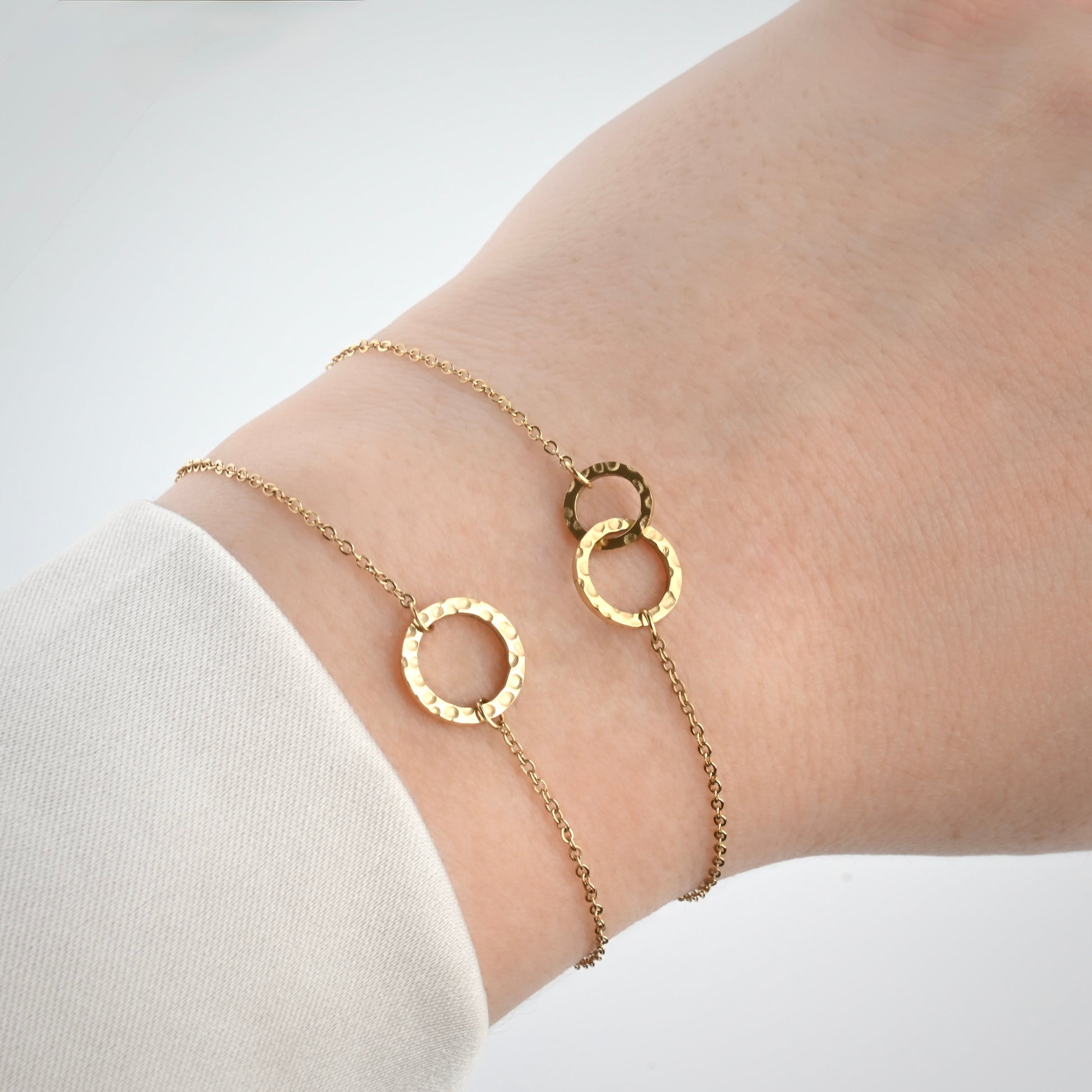 Two gold chain bracelets with hammered circle links on a wrist, one with a single medium circle, one with two different sized interlocking circle charms.