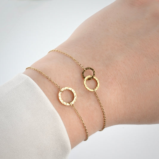 Two gold chain bracelets with hammered circle links on a wrist, one with a single medium circle, one with two different sized interlocking circle charms.