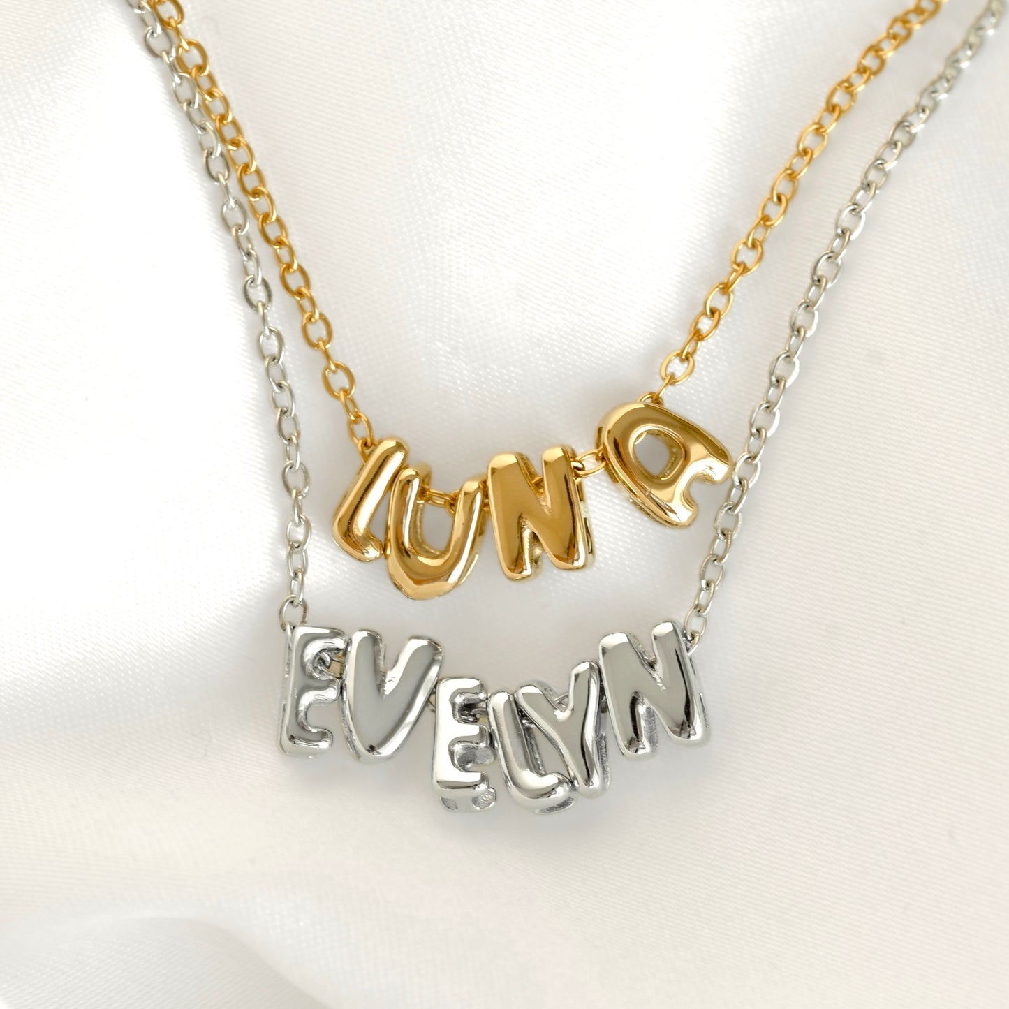 Two name necklaces with 'LUNA' in gold lettering on a gold chain and 'EVELYN' in silver lettering on a silver chain, displayed on a white satin background.