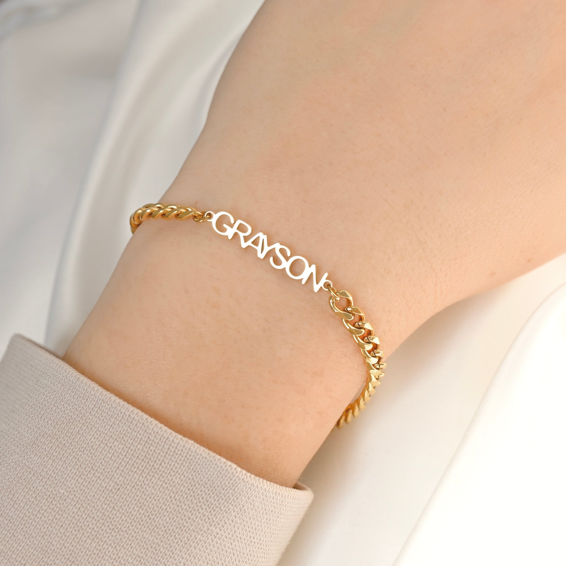 A close-up of a thick gold cuban chain bracelet worn on a person's wrist, featuring the name 'GRAYSON' spelled out in raised letters. The person is wearing a light beige long-sleeve top against a soft white background.