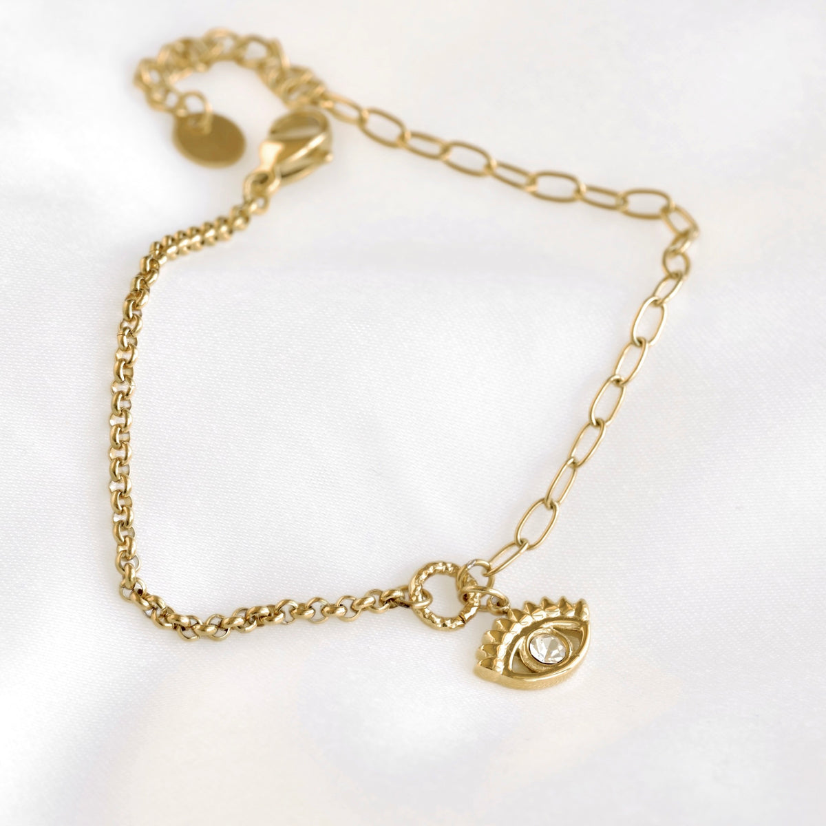 A gold chain anklet featuring a small gold eye charm with a clear rhinestone, displayed on a soft white fabric background.