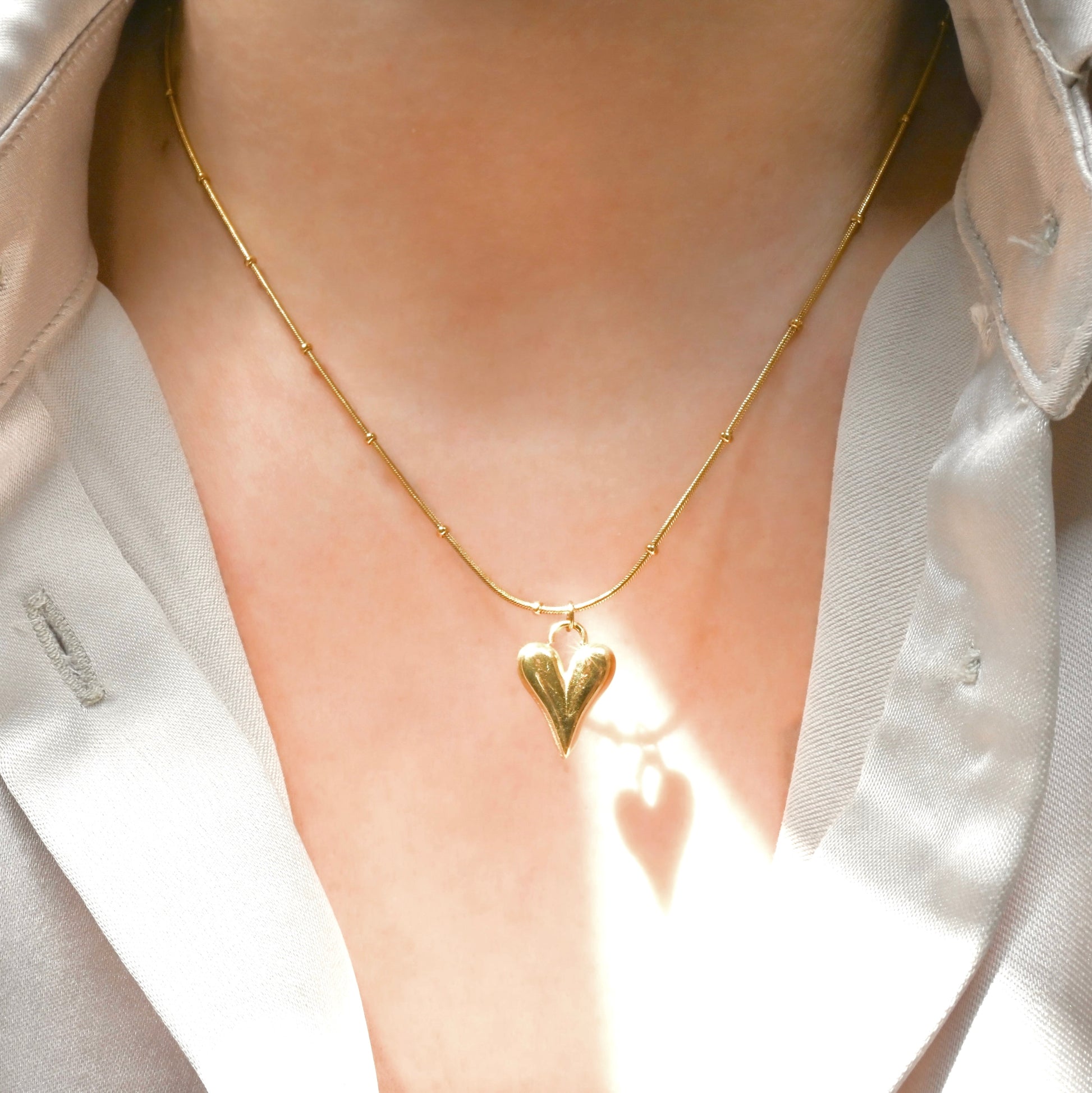 Gold heart necklace worn by a person with a light-colored shirt under direct sunlight.