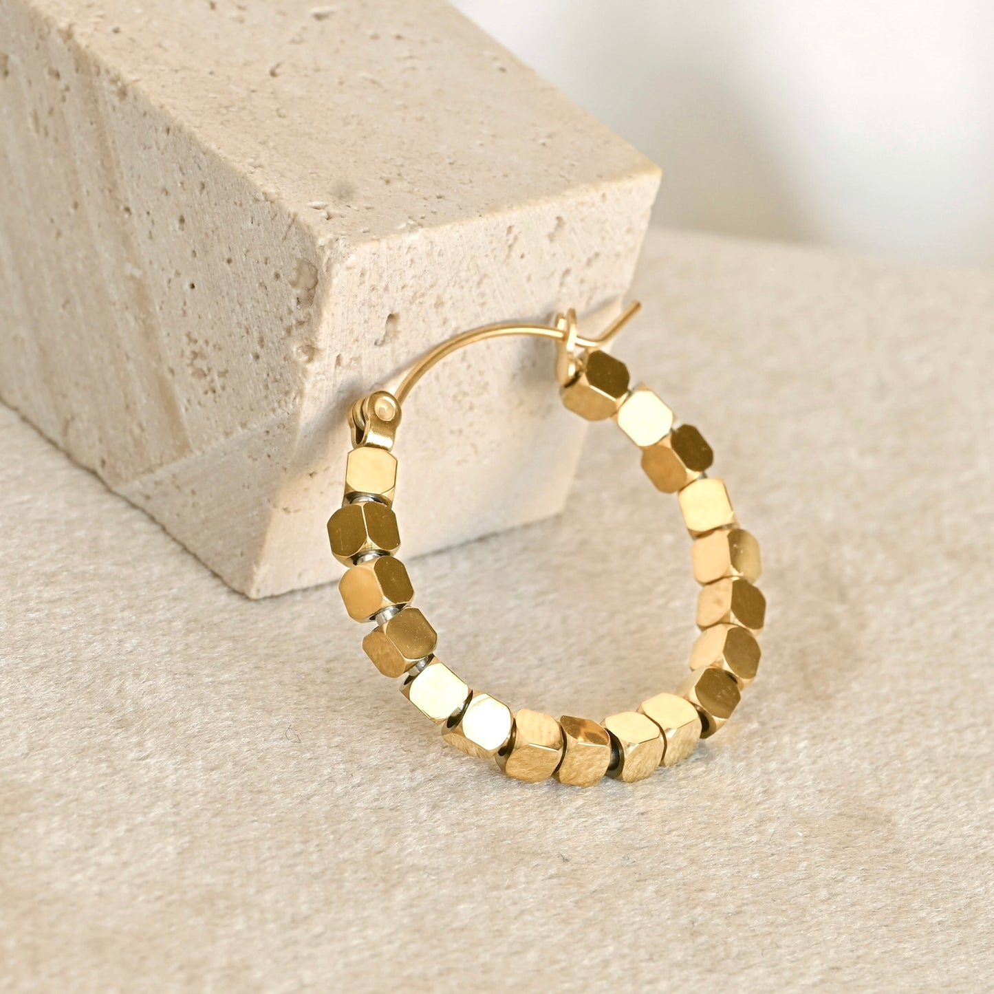 Gold hoop earrings made of cube beads, displayed on a textured beige surface next to a light stone block.
