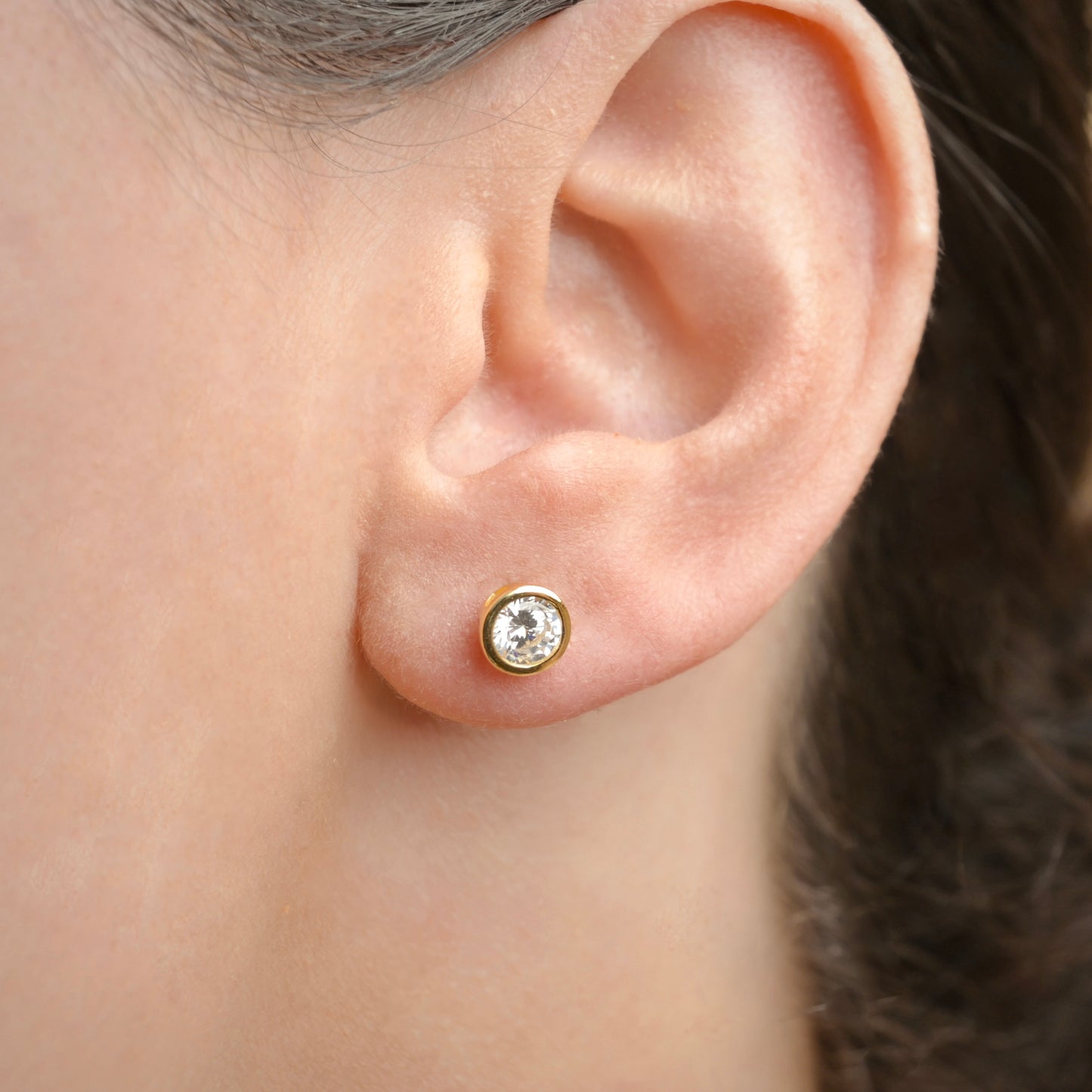 Close-up of an ear wearing a gold  solitaire earring with a clear gemstone.