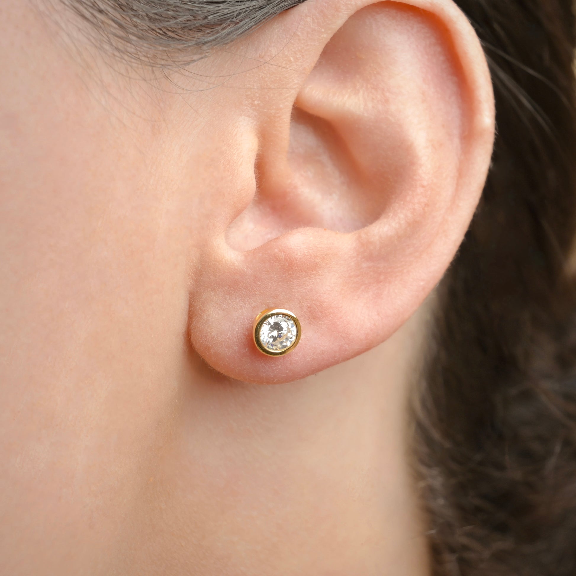 Close-up of an ear wearing a gold  solitaire earring with a clear gemstone.