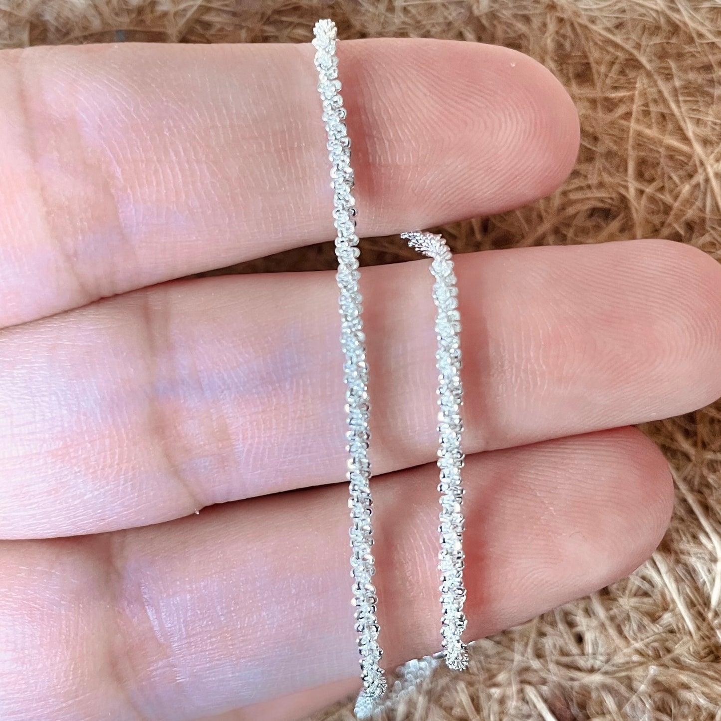 Silver shimmer anklet held between fingers against a natural fiber background