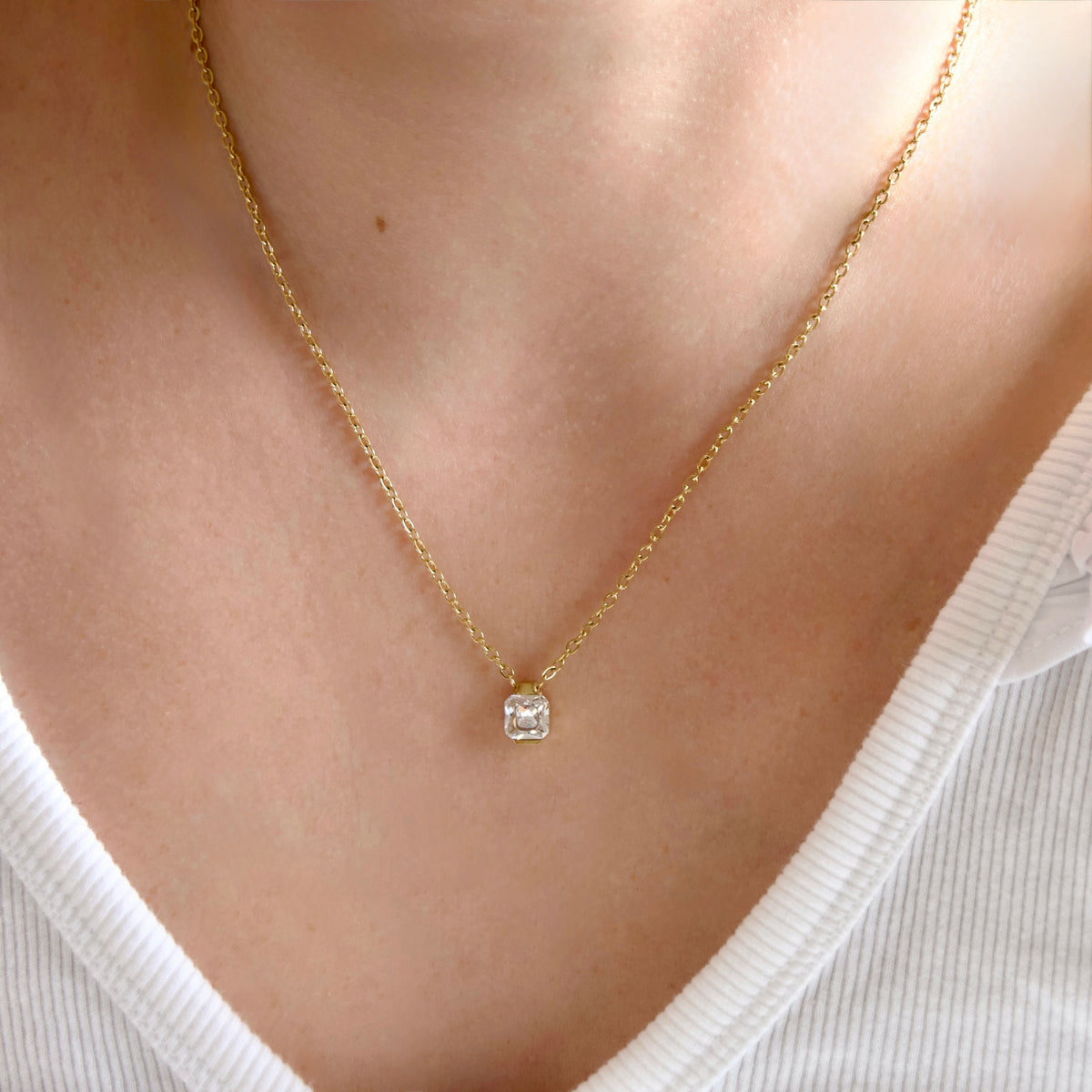 A close-up of a person's neck and chest wearing a delicate gold chain necklace with a small square clear gemstone pendant, paired with a white ribbed V-neck top.