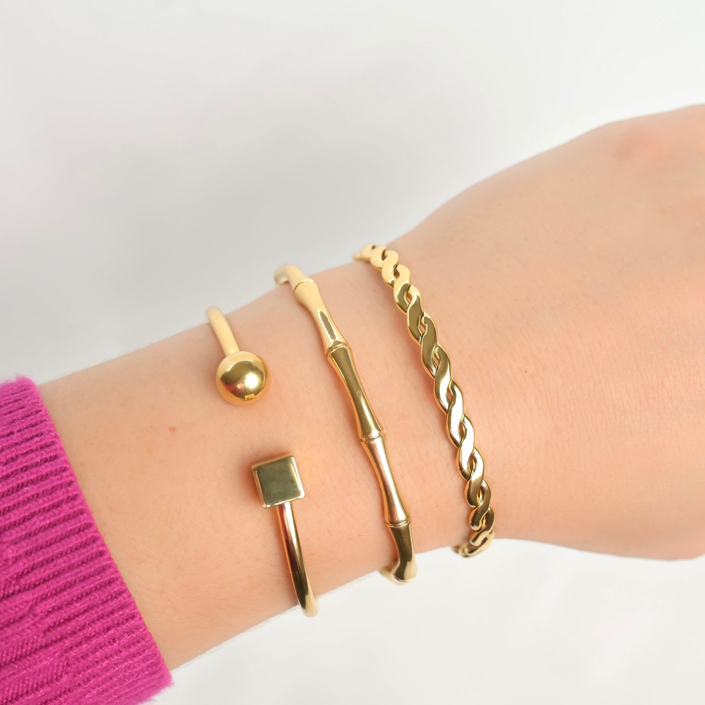Gold thin cuff bracelets including cube&ball, molten and braided designs,  on a wrist with a pink sleeve against a white background