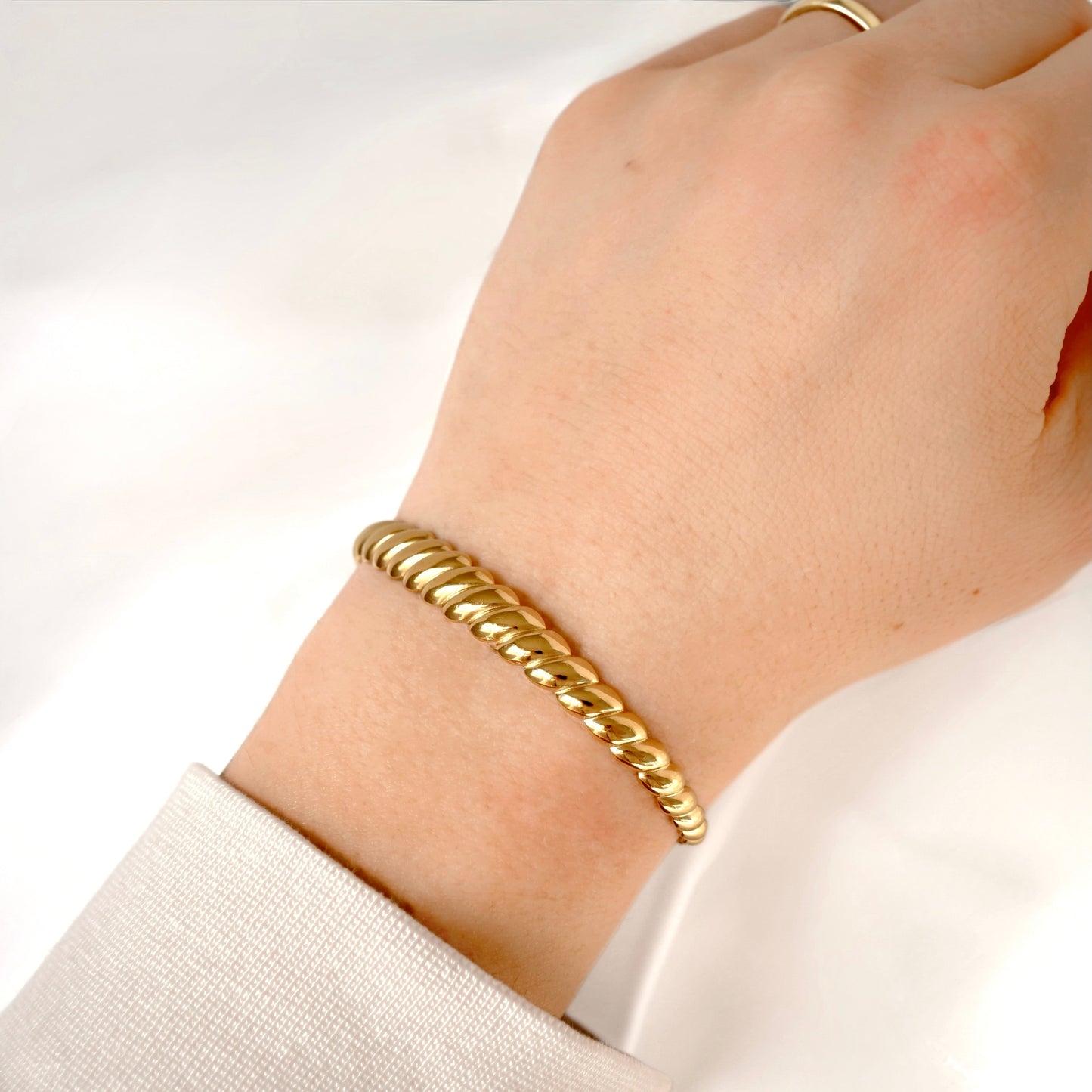 A close-up of a person's wrist wearing a twisted gold rope-style bracelet, with a light-colored sleeve visible at the cuff and a matching gold ring on the finger.