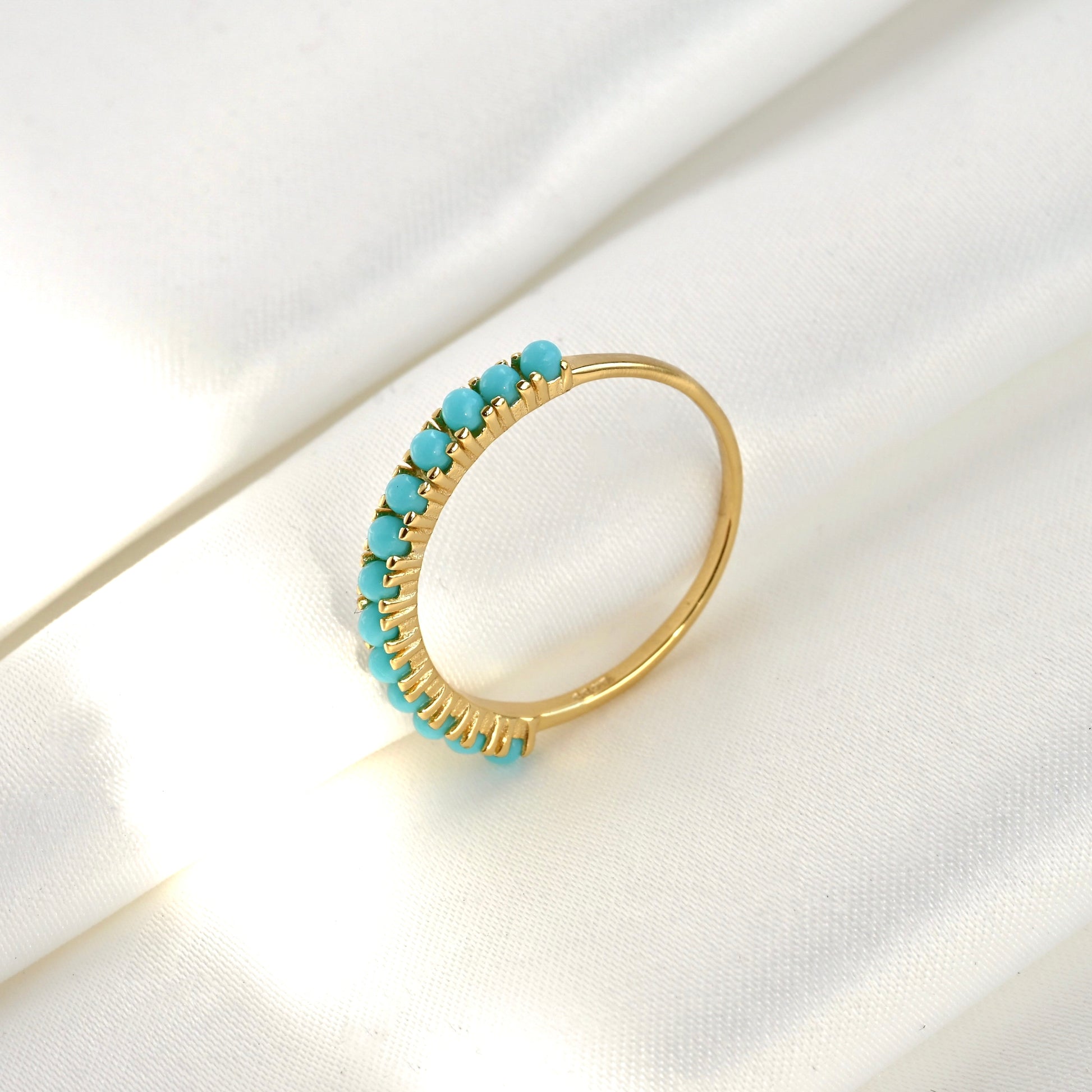 Gold ring with turquoise stones leaning upright against a folded white fabric, the turquoise stones faicng left side.