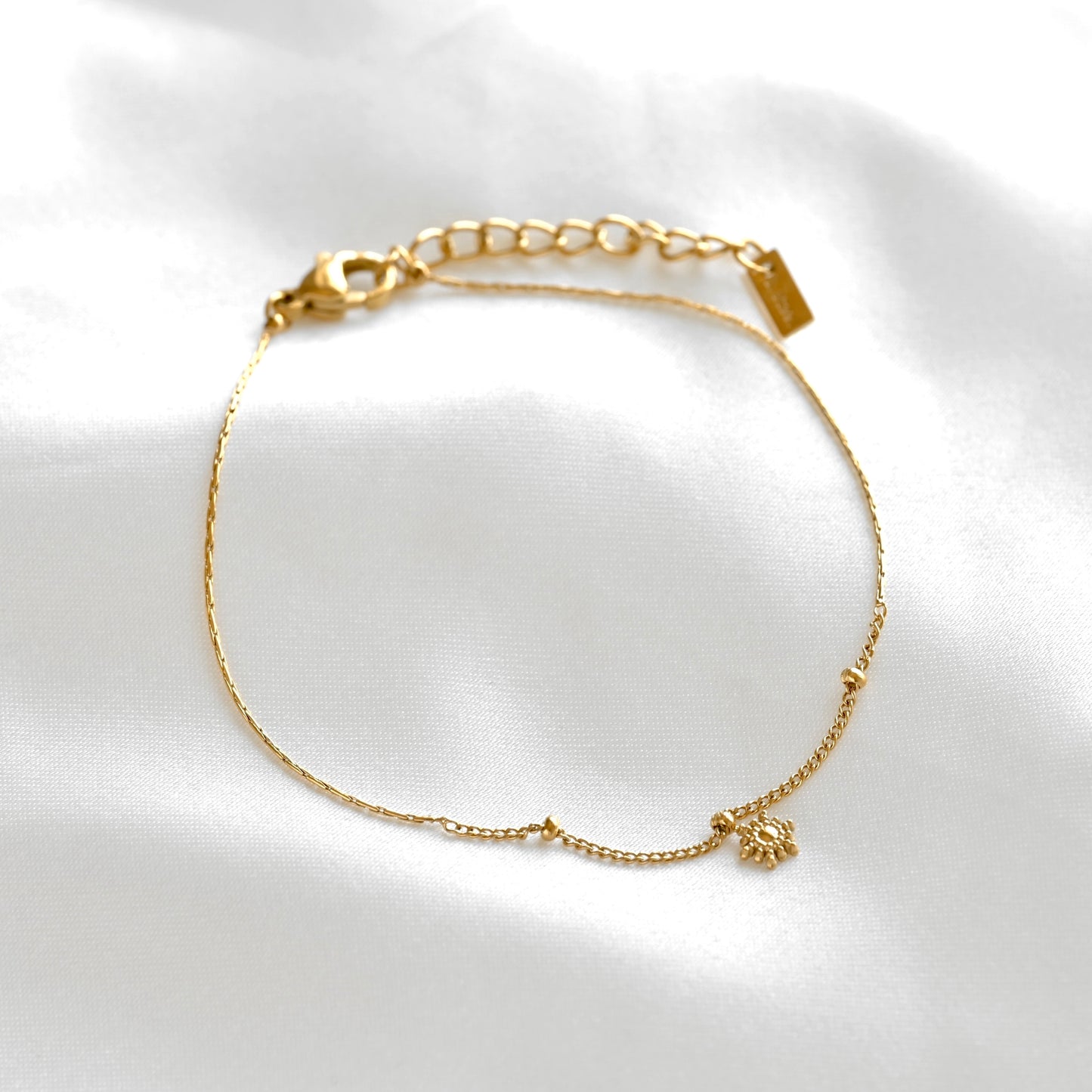 A delicate gold satellite chain bracelet with a tiny sunburst charm, displayed on a soft white satin fabric background.