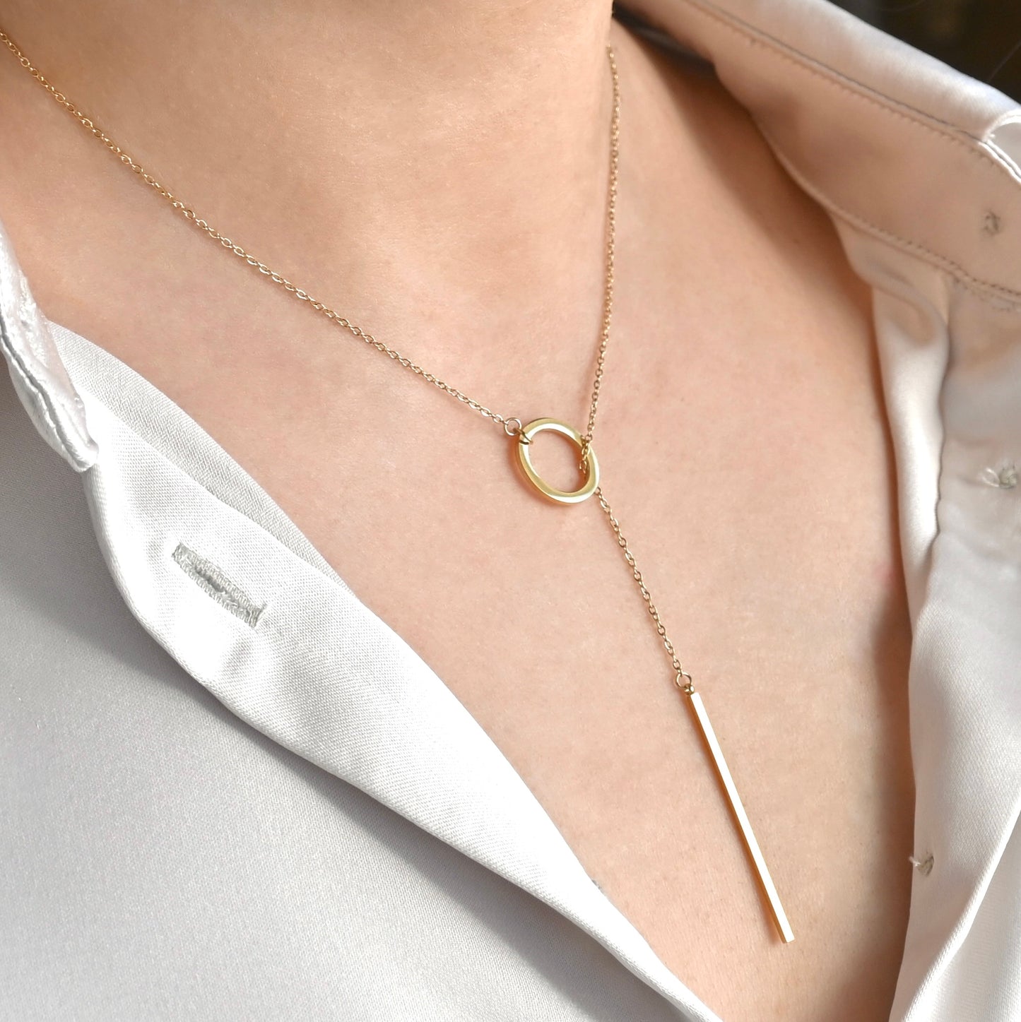 Gold lariat necklace with a circular pendant and a bar pendant worn by a person in a white shirt.