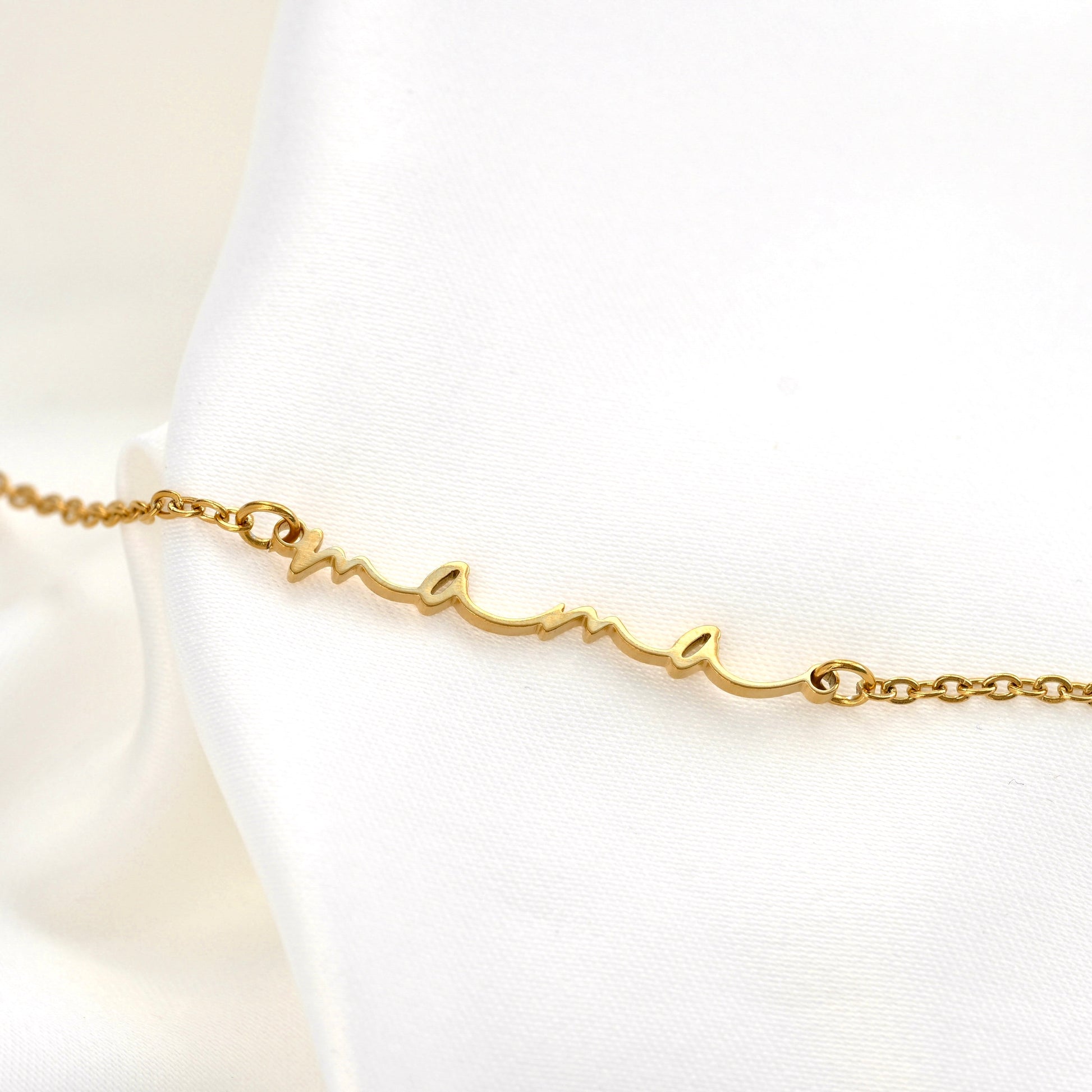 A delicate gold mama bracelet with a cursive script nameplate reading 'mama', displayed on a white satin fabric