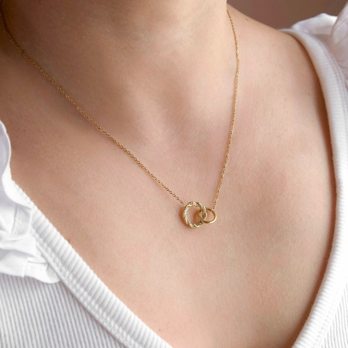 A close-up of a model wearing a delicate gold chain necklace with two interlocking open circle pendants, styled with a white ruffled-shoulder top.