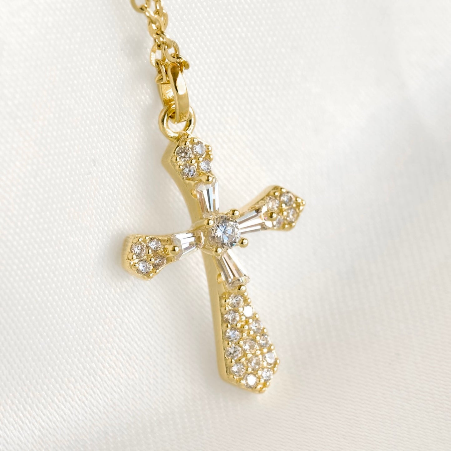 A gold cross pendant necklace adorned with clear crystals, shown on a white textured fabric background.
