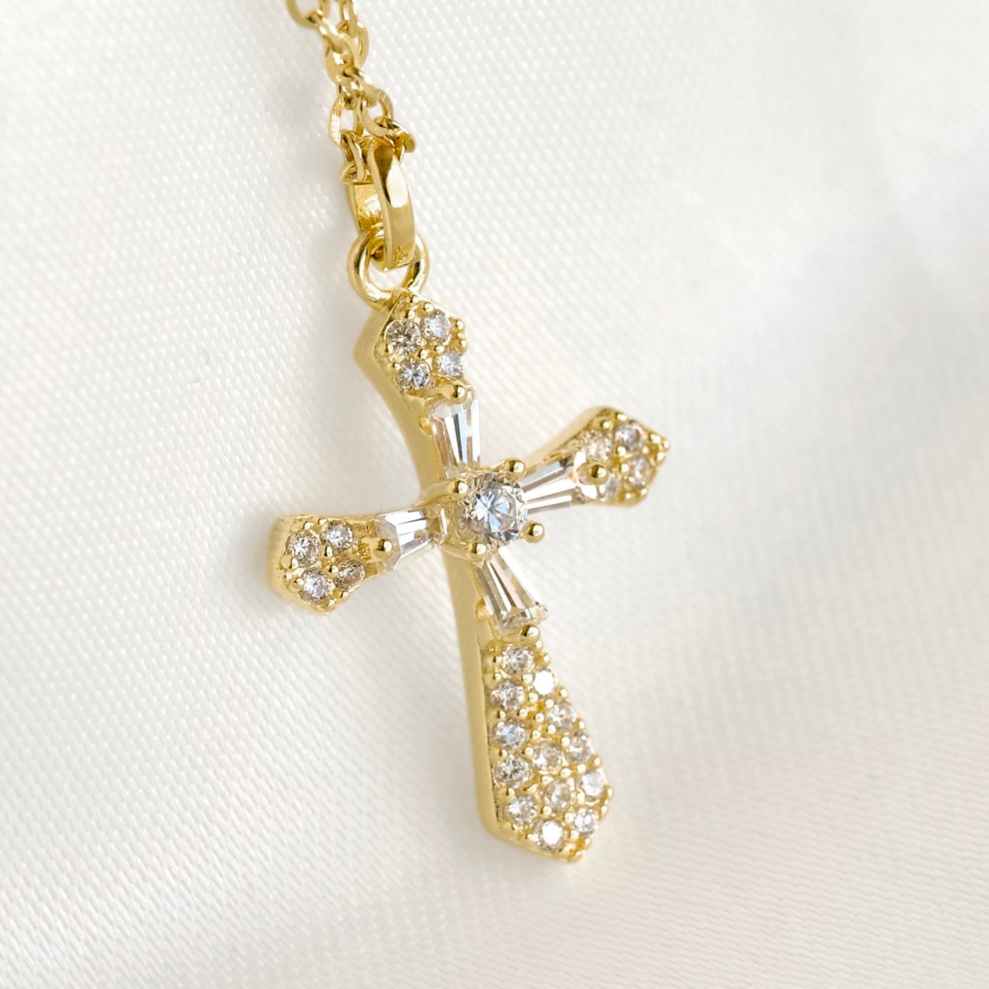 A gold cross pendant necklace adorned with clear crystals, shown on a white textured fabric background.
