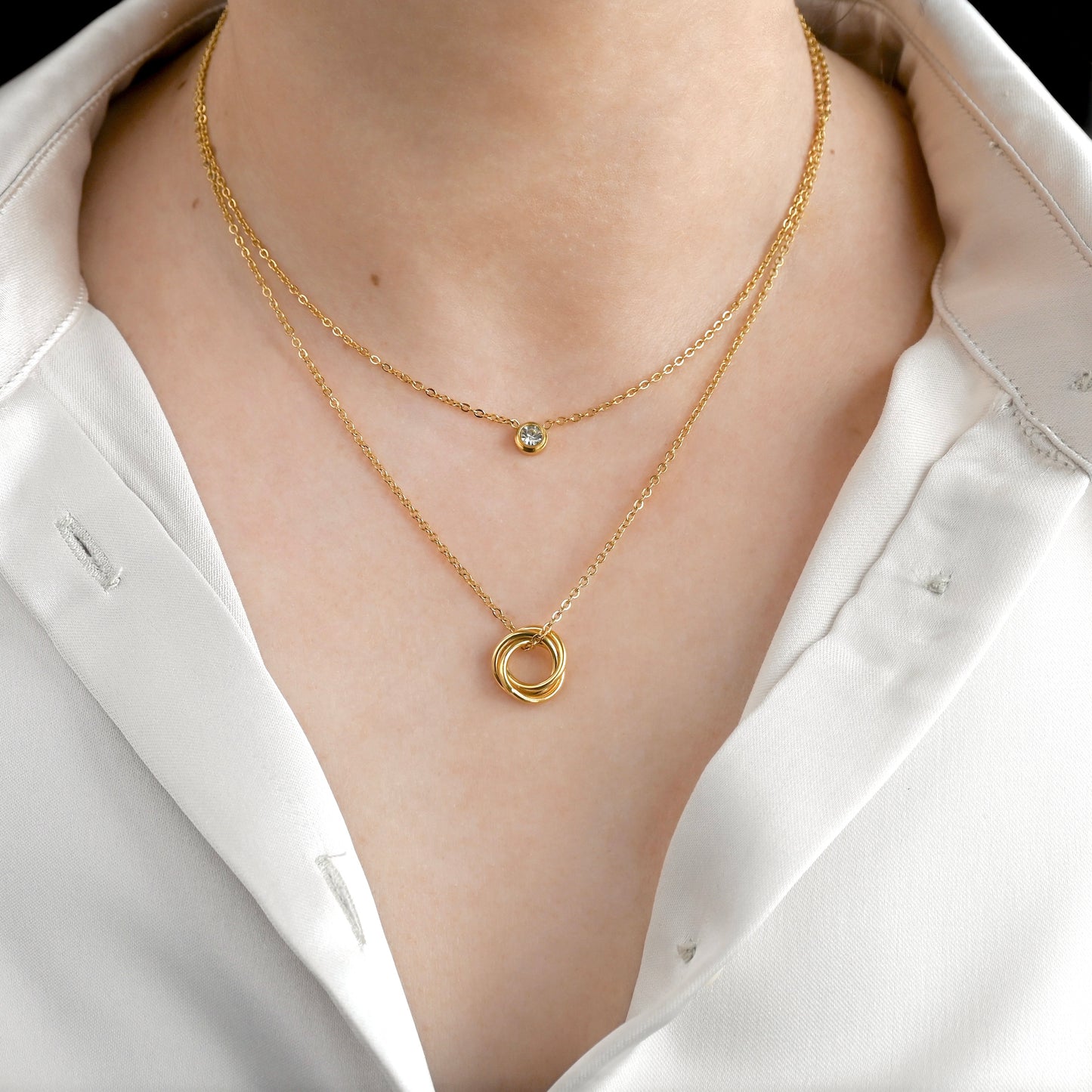 Front view of gold necklace set with a trinity knot pendant and small bezel stone worn by a person in a white shirt.