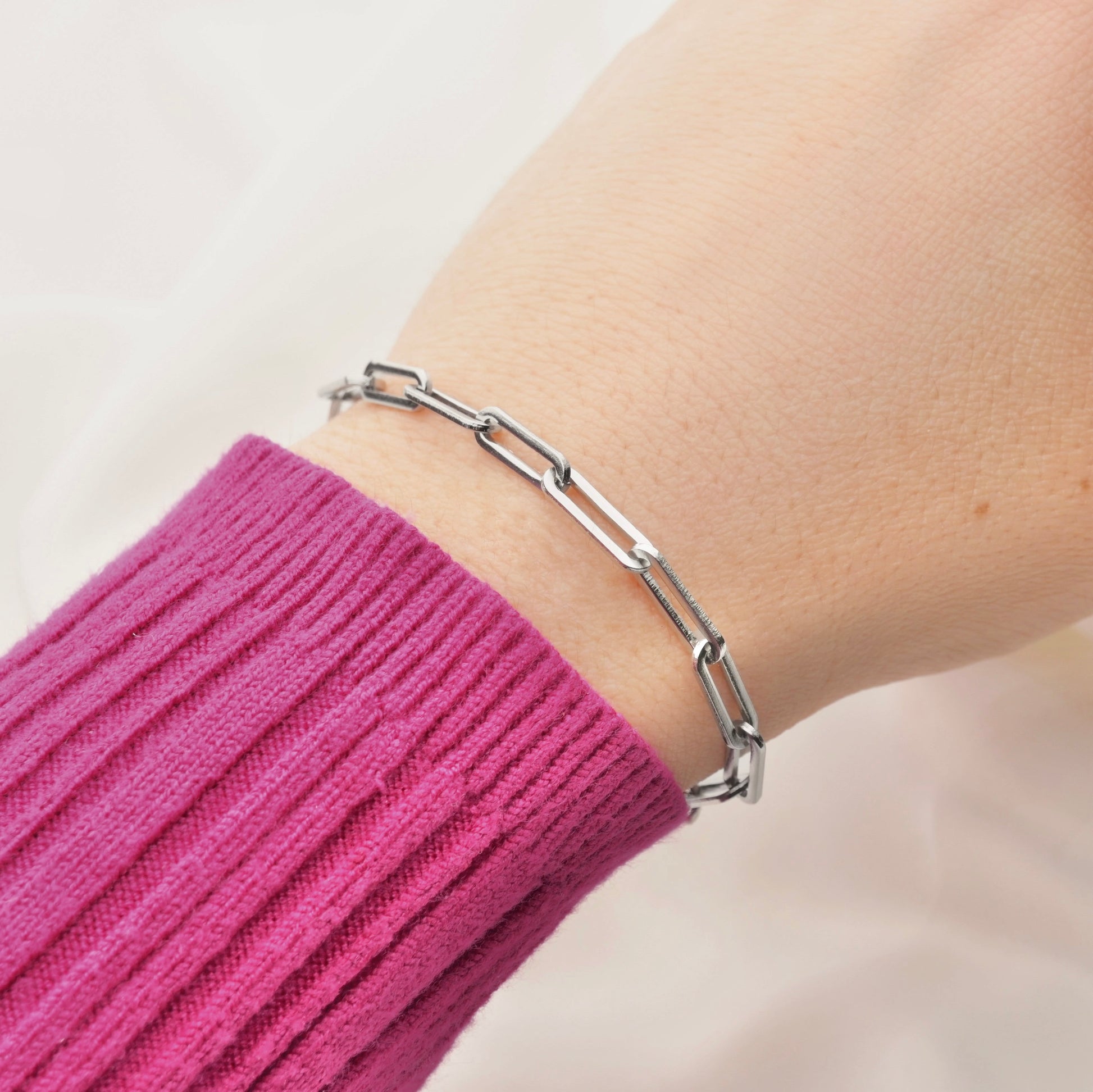 Silver rectangular link chain bracelet on a wrist with a pink sleeve against a neutral background