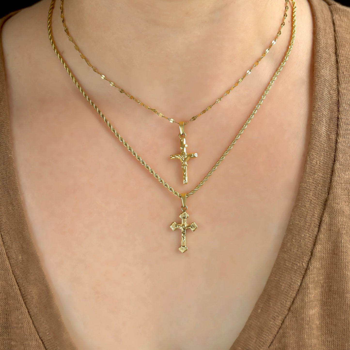 Two gold cross necklaces with different chains and pendants layered  on a person wearing a brown top.