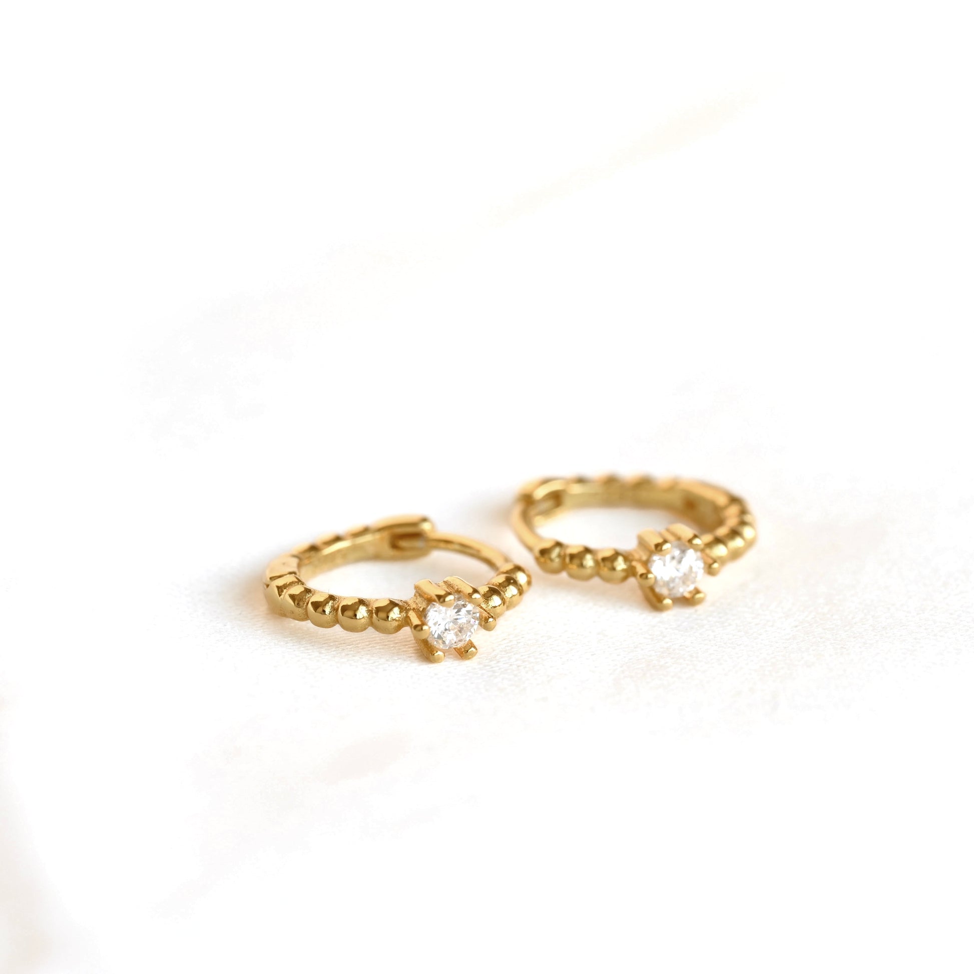 A pair of small gold huggie hoop earrings, each featuring a central clear round gemstone in a prong setting, resting on a white surface.