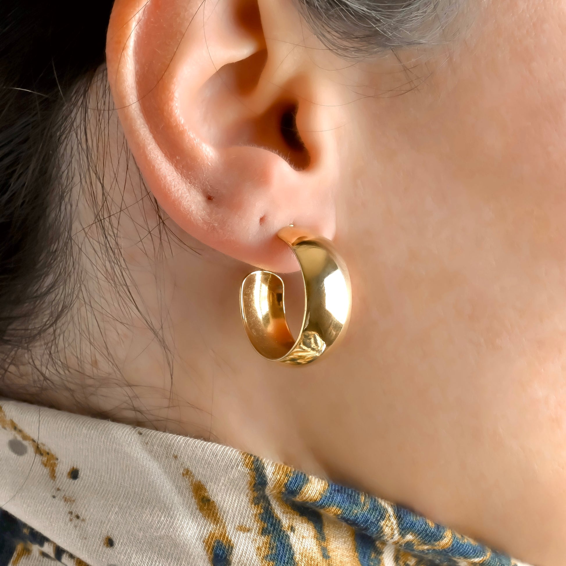 Gold chunky dome hoop earrings worn by a person with a patterned fabric background