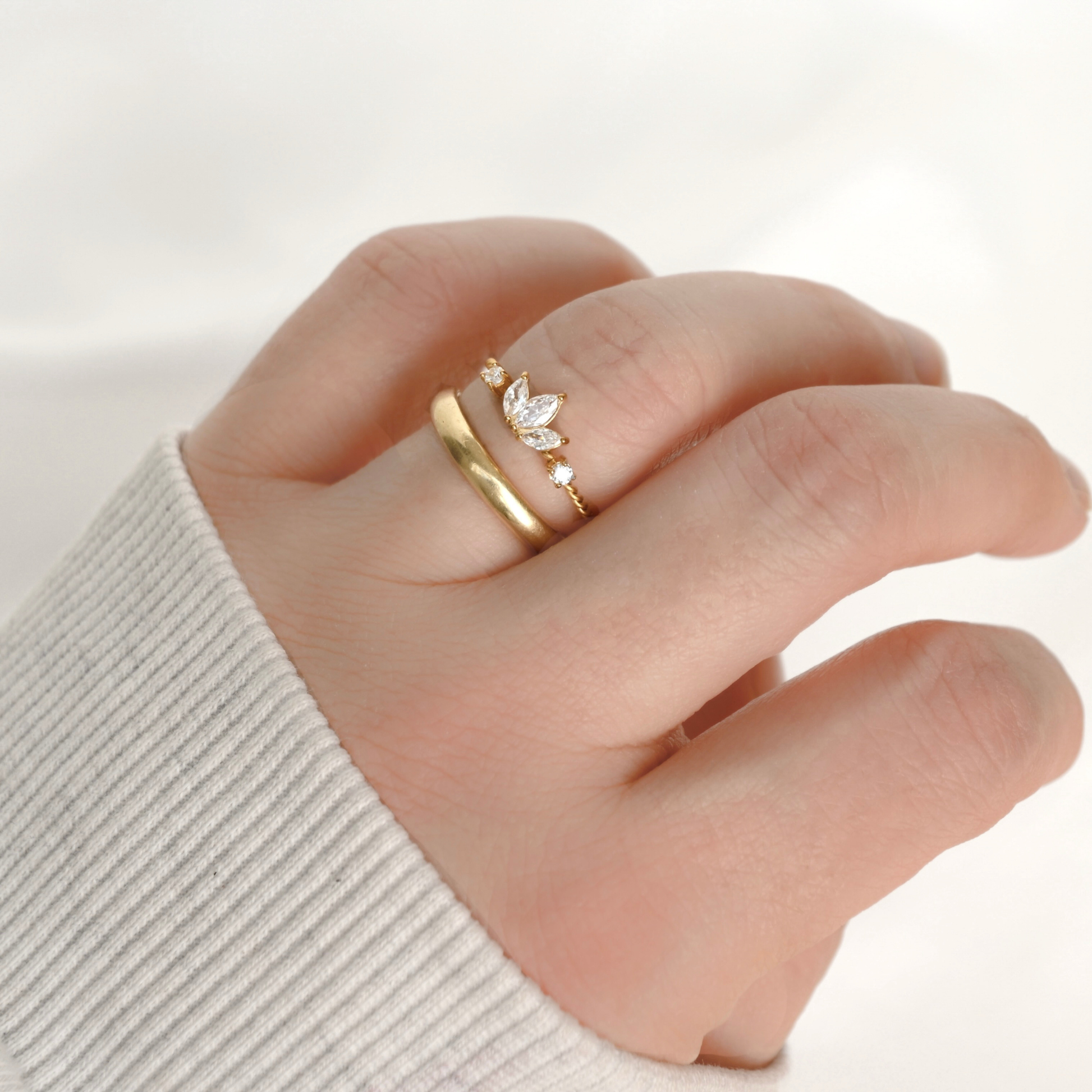 A close-up of a hand wearing two gold rings, including one delicate ring with a three cz clusters forming a lotus flower on a delicate twisted band against a soft white background.