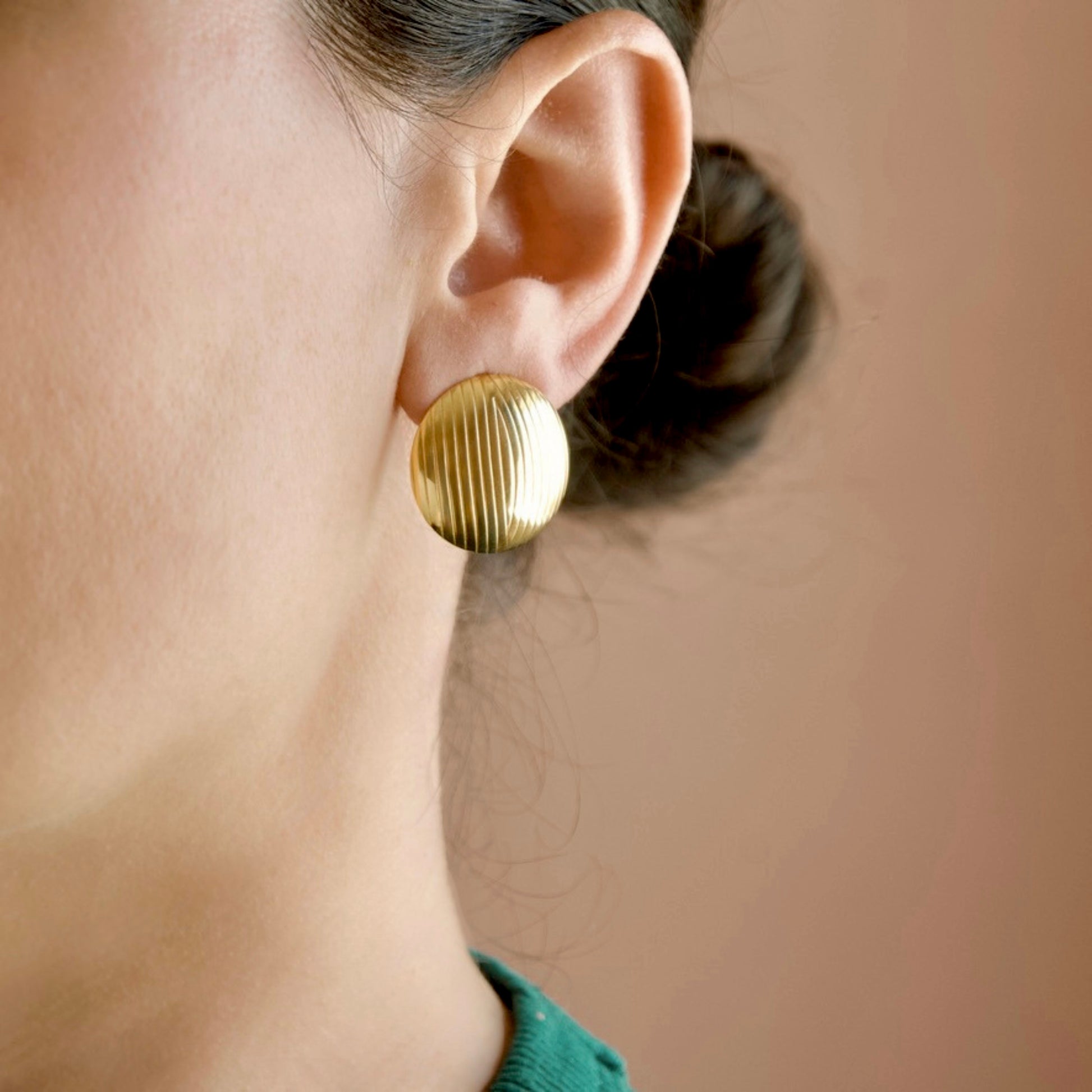 A close-up side profile of a person wearing a single round gold earring with vertical ridges, set against a soft warm beige background.