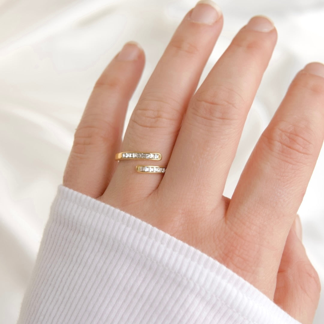 A close-up of a hand wearing two slim gold rings with small clear stones, set against a soft white fabric background.